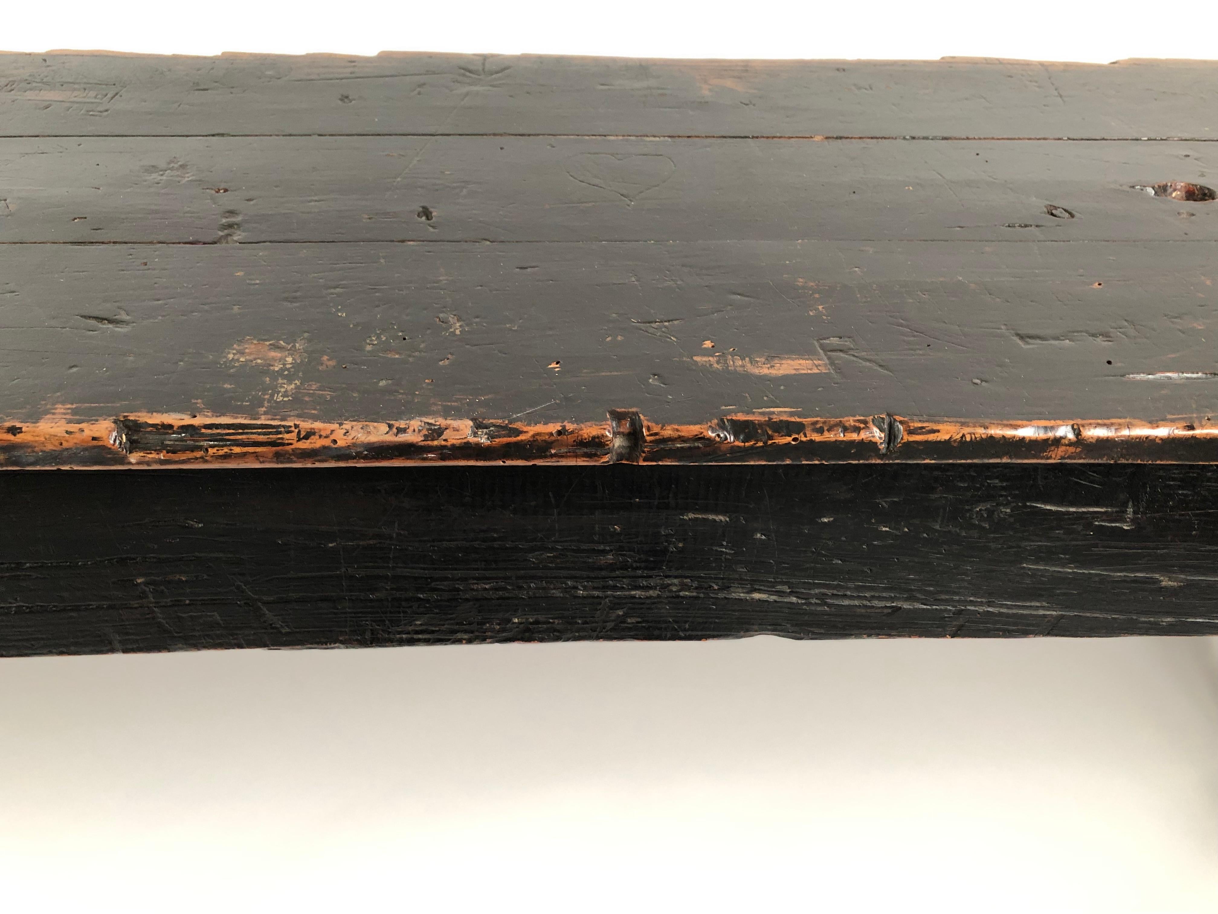 An unusually long narrow country console table in old, weathered black paint, the rectangular top carved with graffiti, over four square section legs joined by cross stretchers on each end. This table would work very well as a sofa table, serving