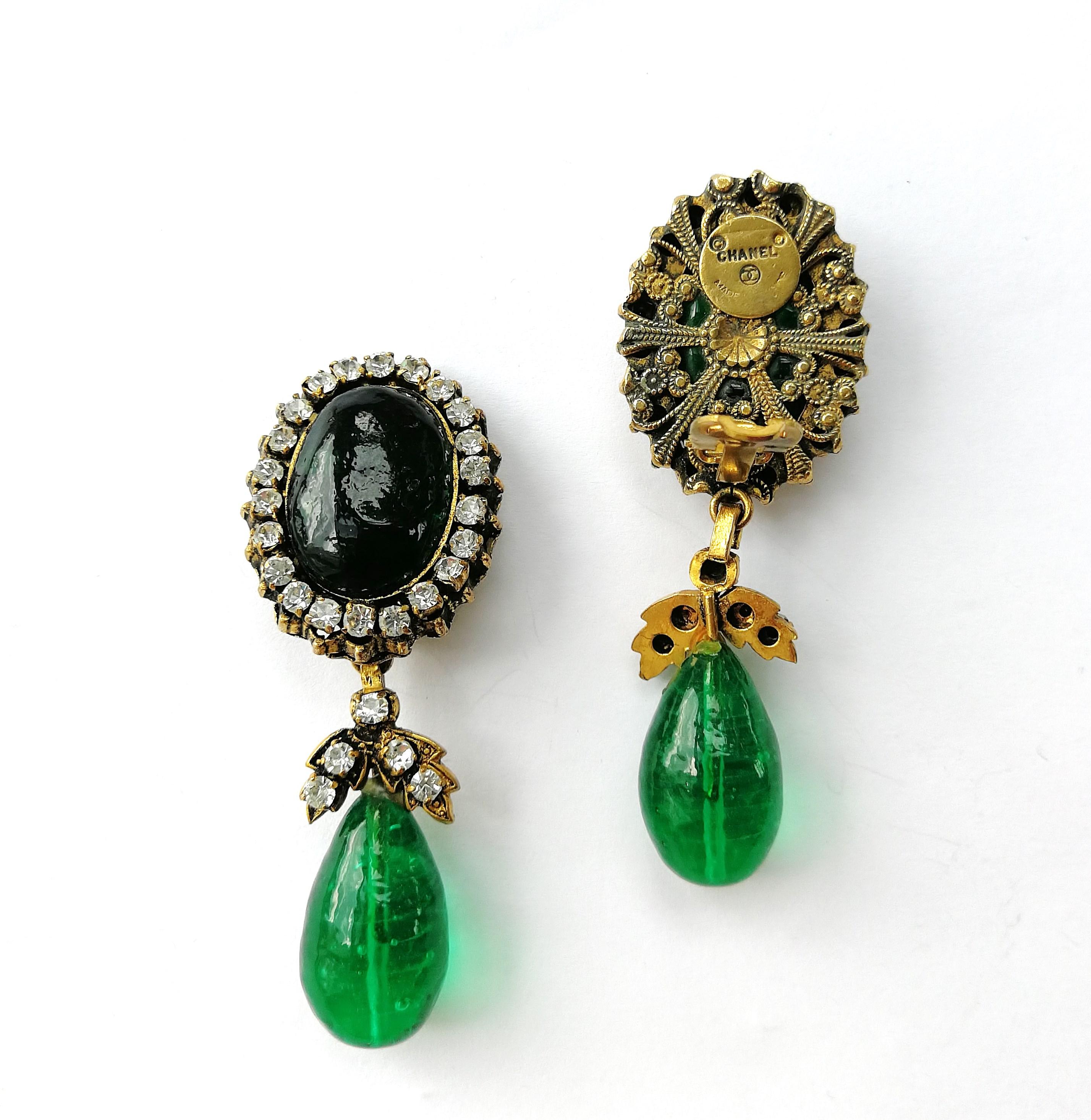 A beautiful and eye catching pair of emerald and clear paste drop earrings, made by Maison Gripoix for Chanel in the late 1970s/early 1980s. A large emerald poured glass cabuchon sits on the ear, circled in clear pastes, and a luscious emerald