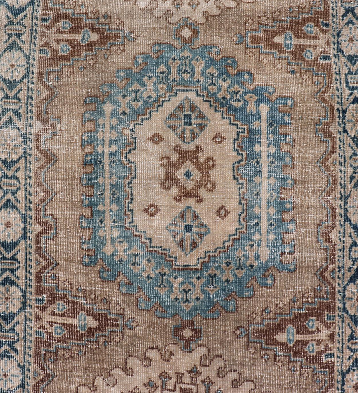 This antique Persian Heriz runner features a Sub-geometric medallion design rendered in blue, tan, brown and cream tones, set upon a tan background. A complementary, multi-tiered border encompasses the entirety of the piece; making it a marvelous