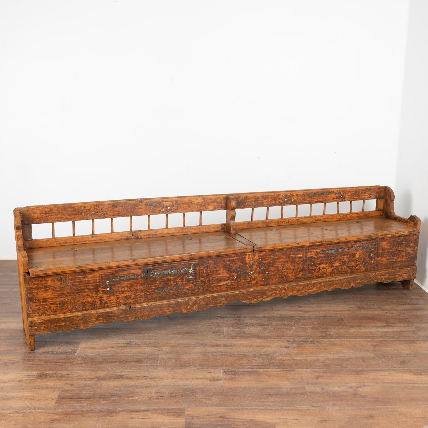 Antique hand painted bench with divided storage at over 9' long .
Traditional Eastern European folk art hand-painting reveals faded flourishes/flower borders along front against brown painted background. 
Two bench seats open to reveal divided