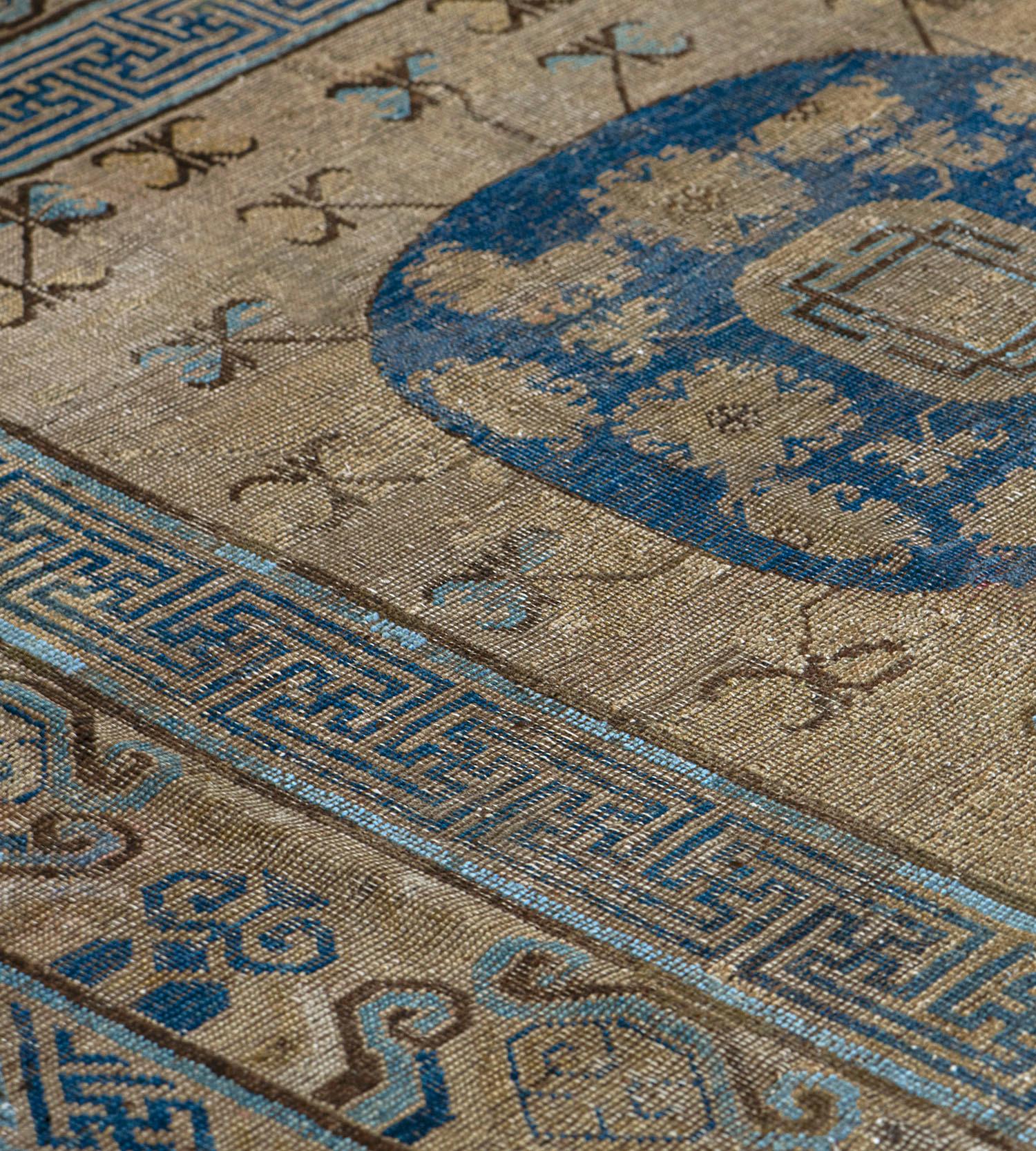 This antique Khotan rug has a dusty sandy-brown field scattered with delicate light blue and golden-yellow delicate flowerhead stems around a column of three royal-blue roundels each containing angular flowerheads and floral vine around a central