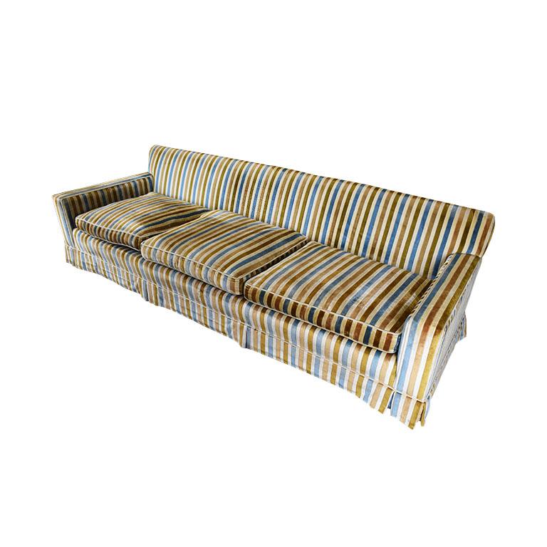 A long vintage striped sofa couch in light blue, gold, yellow, and cream. Created from velvet, this midcentury piece has three removable cushions, track arms, and a skirt. The maker is unknown. Some damage to one of the legs, however structurally