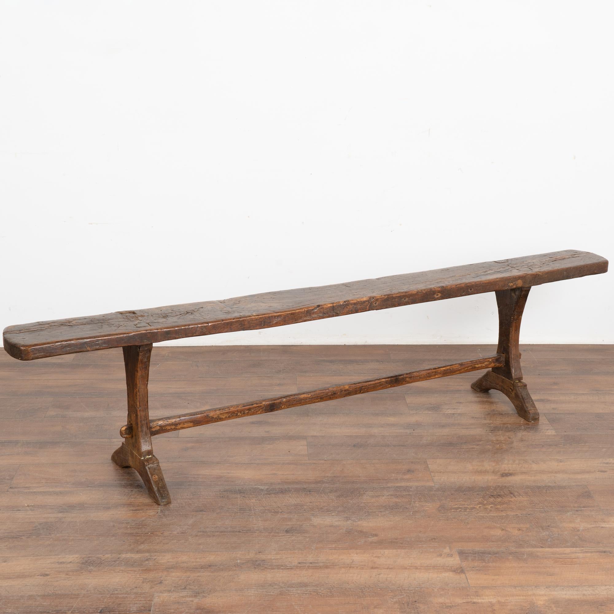 Rustic dark oak wood plank top bench with carved trestle legs and long stretcher.
The seat reveals generations of use with nicks, cracks, gnarls and gouges which add character to the narrow bench. 
Restored and waxed, this bench is just over 6.5'
