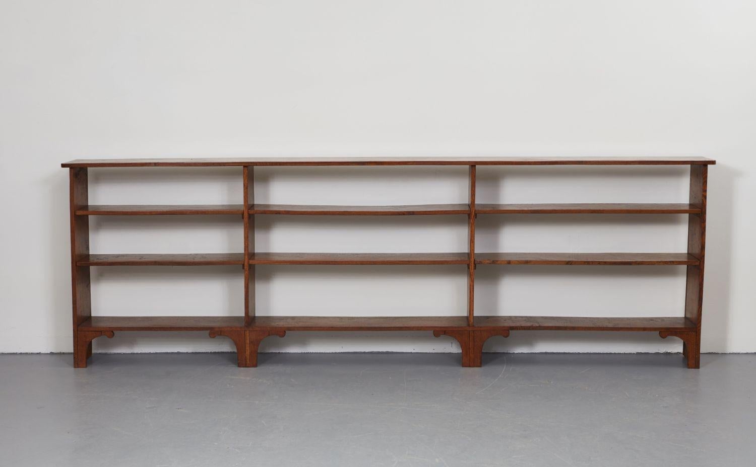 A long open bookcase or storage unit with nine open bays and a top surface shelf. Supported on raised feet with shaped corner brackets. In solid oak with good graining and color. Useful narrow proportions for use under a window or as shoe and bag