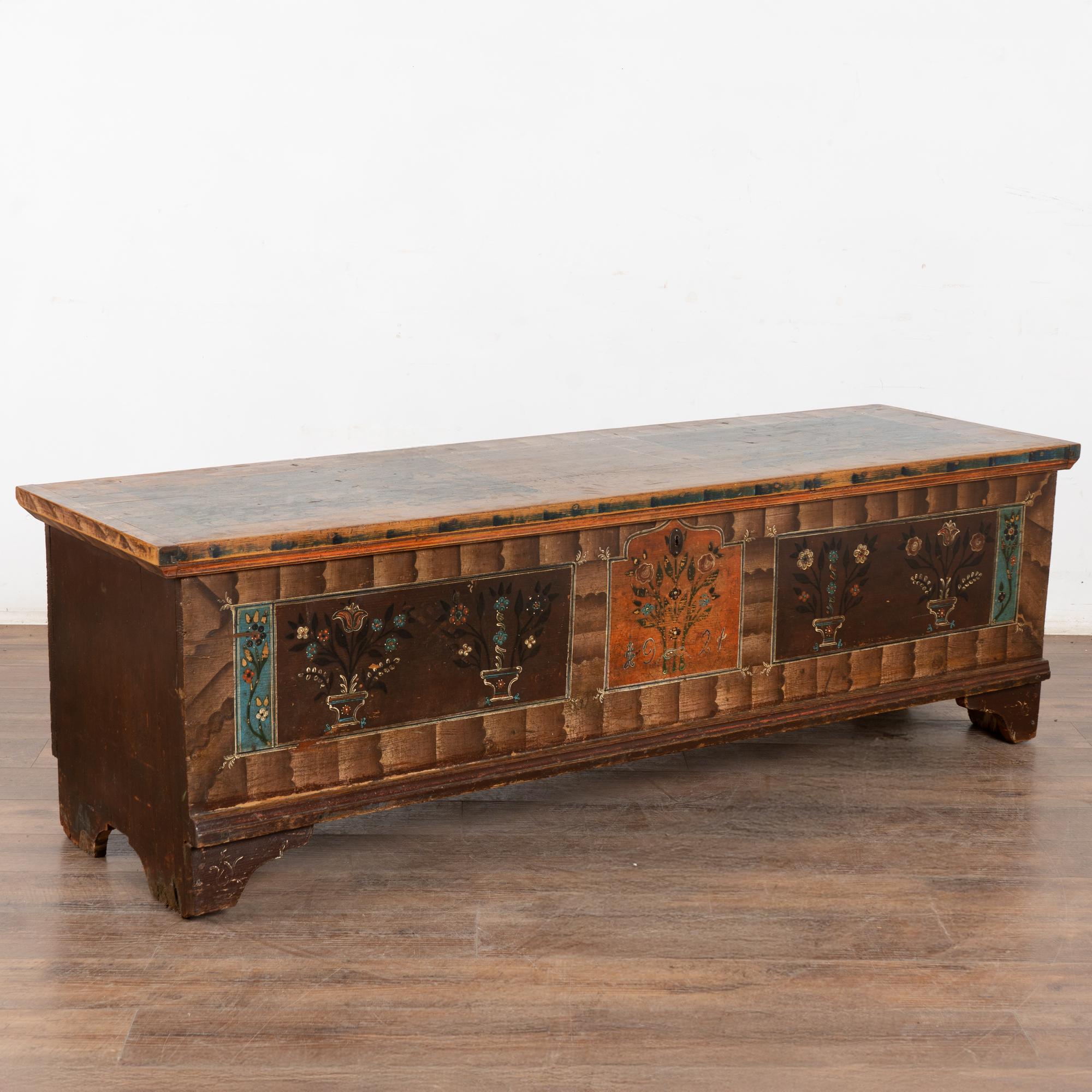 The painted finish is all original on this rustic pine flat top narrow trunk which may also serve as a bench. Flowers were a traditional embellishment as seen on the front of this handcrafted trunk with multi colored flowers contrasting against the