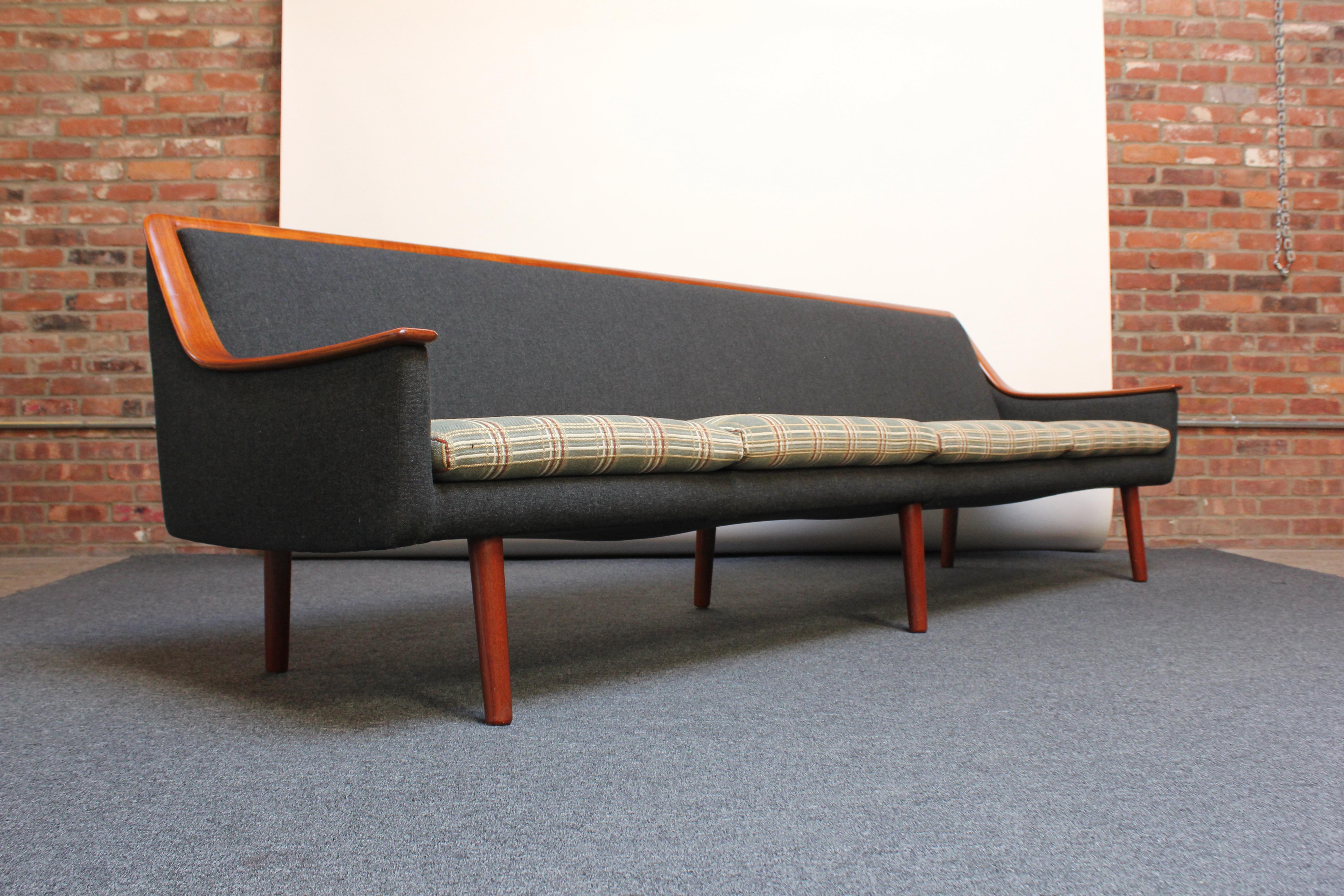 Mid-Century Modern sofa with a continuous, exposed teak arm / back trim (Norway, ca. 1960s). Retains the original upholstery composed of a charcoal wool frame with a contrasting cushion in red, green, white, and oatmeal plaid. The foam and straps