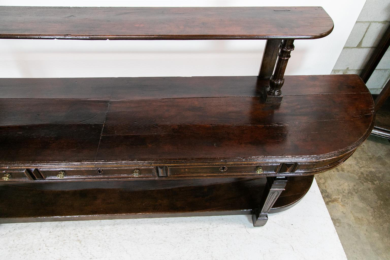 This oak long French server/buffet has an upper shelf supported by carved columns in the front and a flat support in the rear with an exposed mortise and tenon on the top of the shelf. There are three drawers that are framed with applied moldings
