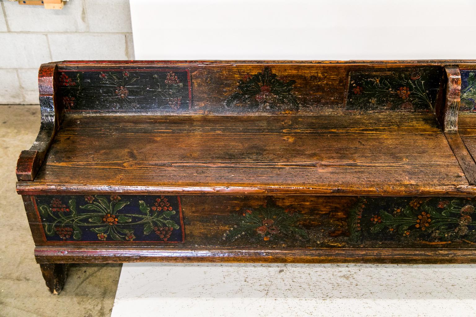 This long painted European bench has floral decoration on the front and backrest. The paint is muted with some wear commensurate with age and use. The backrest is supported by thick shaped brackets on the ends and center. The seats lift up to expose
