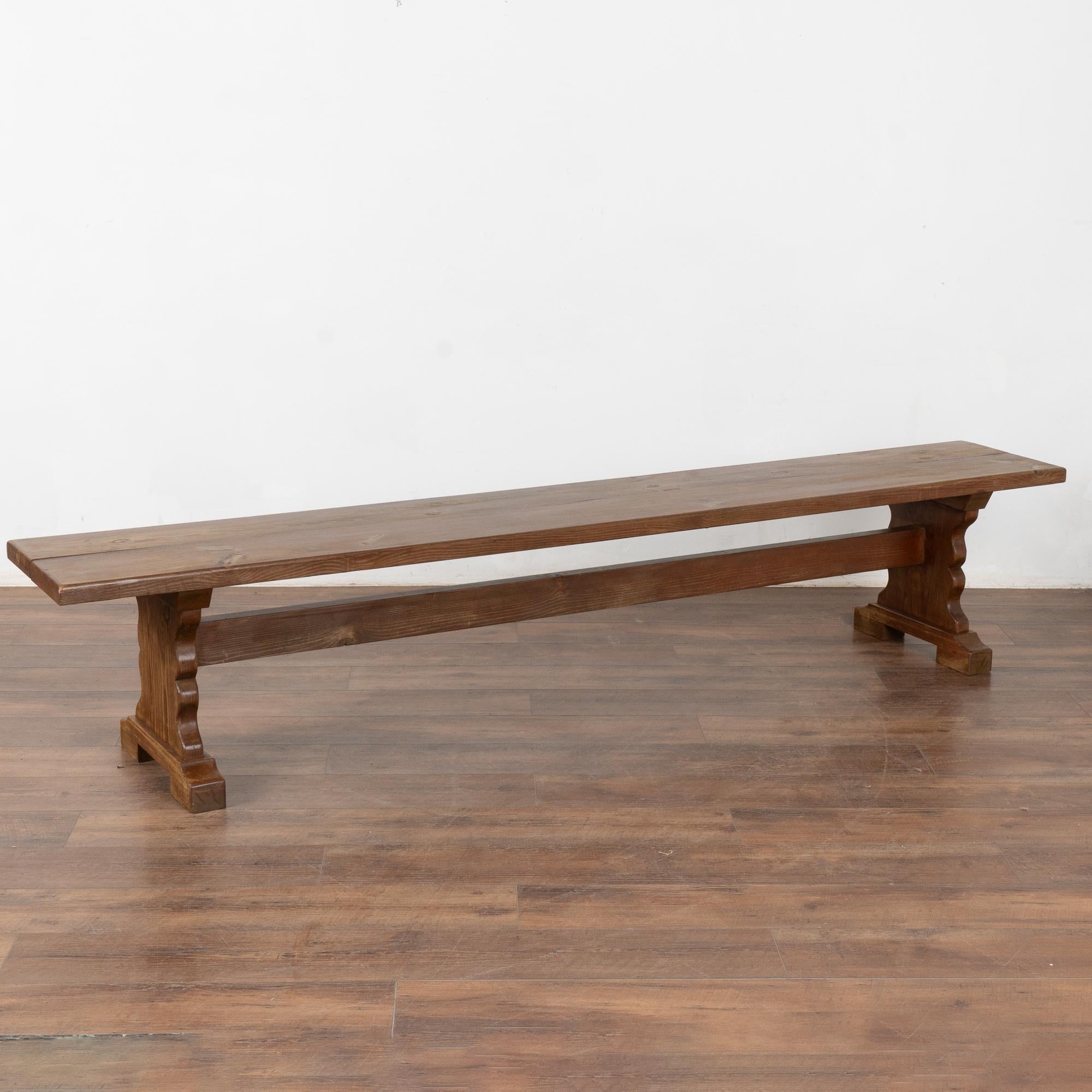 Rustic plank top pine bench with dark aged patina, just under 8' long.
The top reveals generations of use with nicks, old cracks, age related separation of planks and scuffs which add character to the narrow trestle base bench.
Restored, this bench
