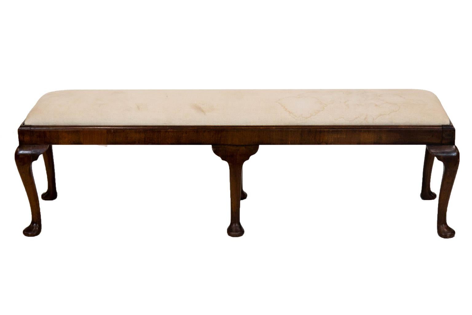 This walnut bench has a carved molded top edge with cabriole legs that terminate in pad feet. The current upholstery has stains.