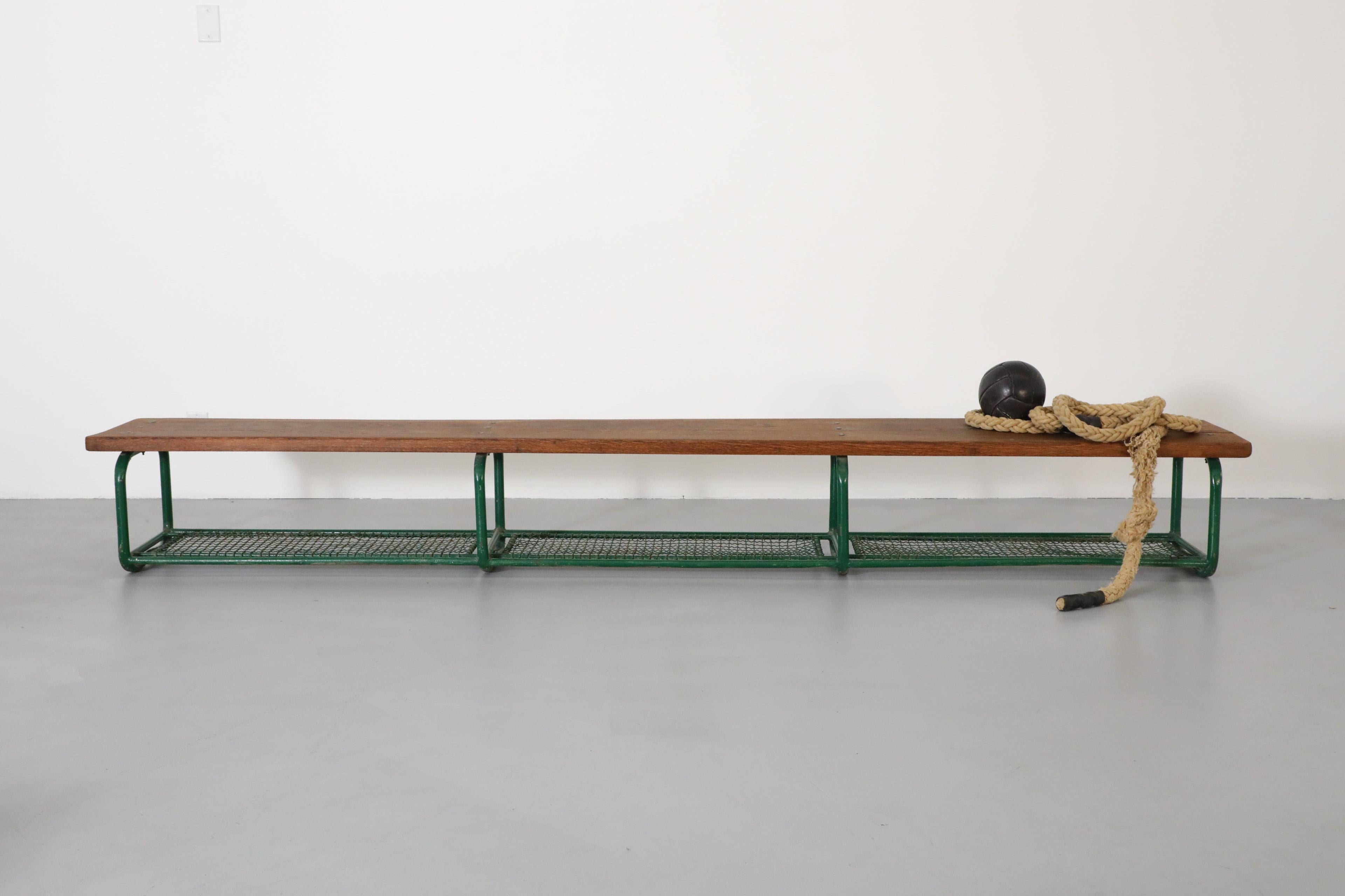 Vintage gymnasium bench with green enameled metal legs and lower basket shelf and solid oak dark stained top. Originally used to house soccer balls and sports equipment. In original condition with visible wear consistent with its age and use.