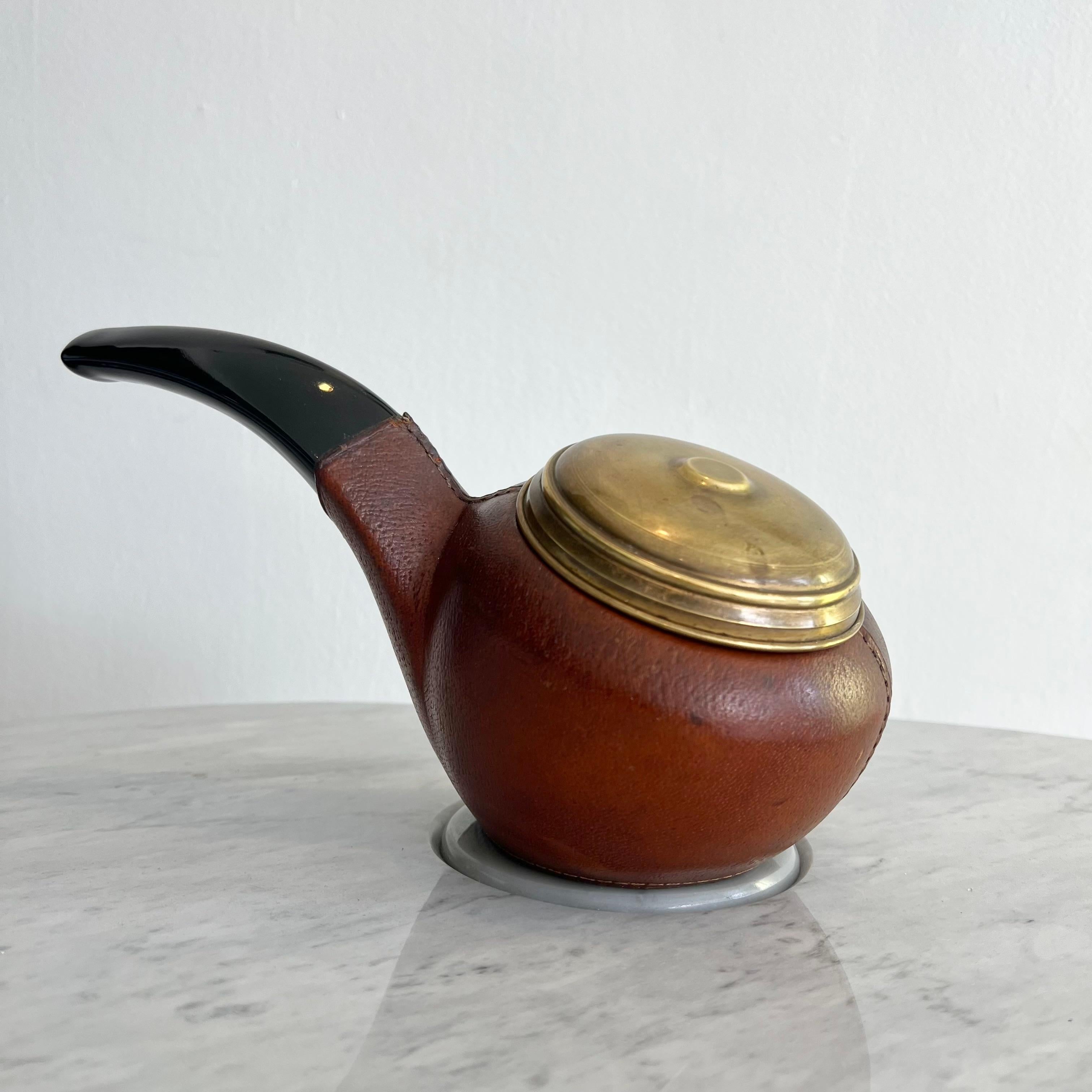 A  rare mid-century cigarette holder in the whimsical form of a smoking pipe. Crafted with a rich brown leather wrapped body, glossy black handle, and a warm brass lid that lifts to reveal a golden cigarette cluster interior. The top opens smoothly,