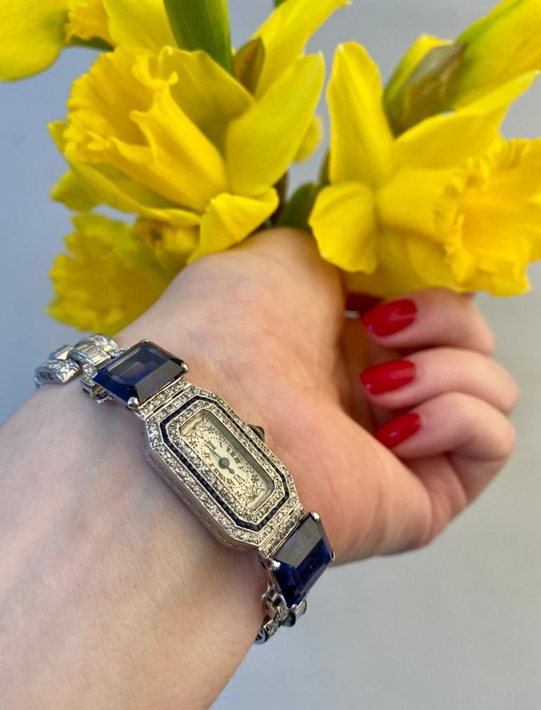 Reloj de pulsera de época, estilo Art Déco, con esfera rectangular de color blanco roto con esquinas recortadas, números romanos negros, manecillas y motivos florales, dentro de una caja engastada con diamantes y zafiro sintético, la corona