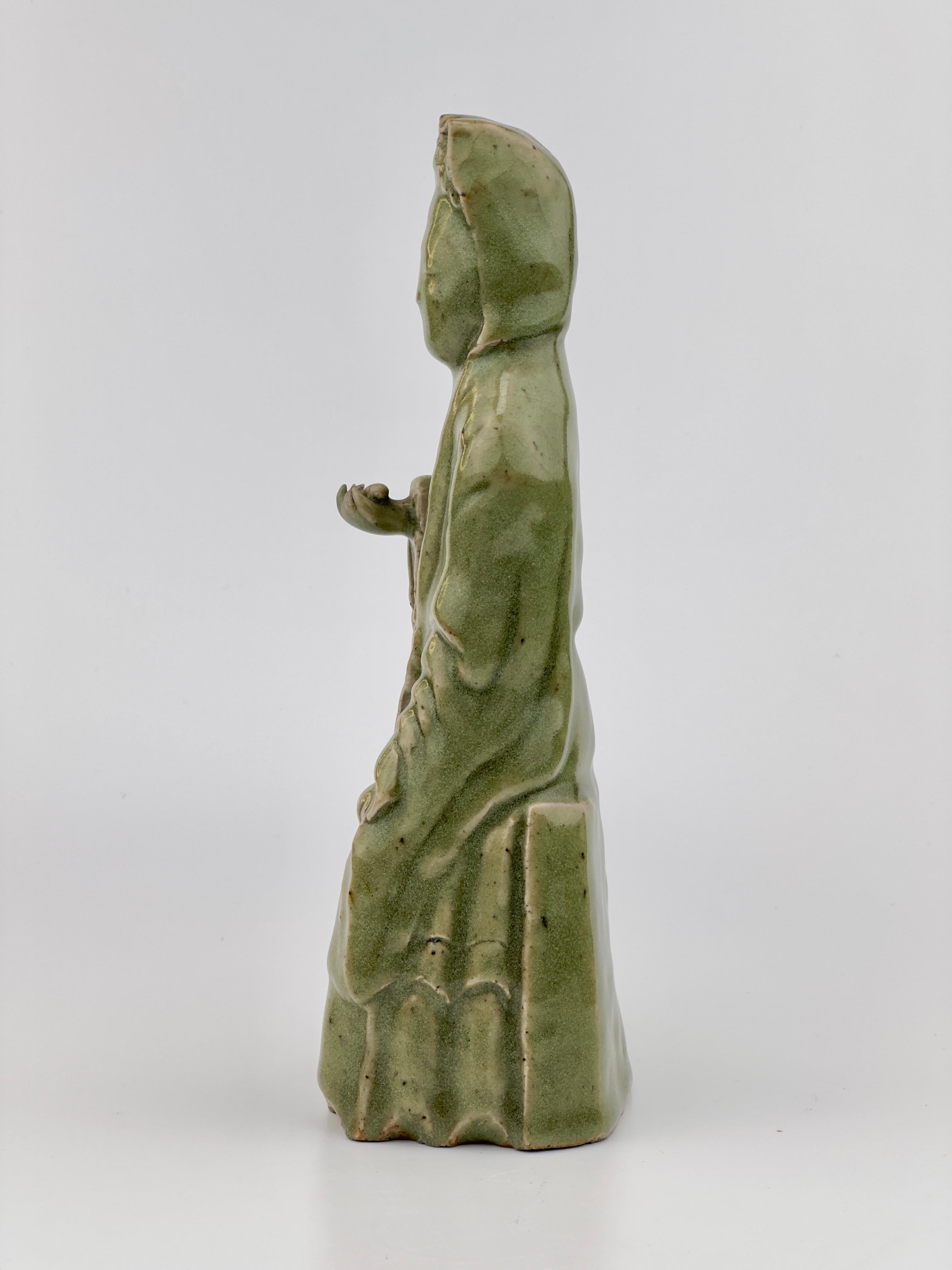 This sculpture is a Longquan celadon from the Ming Dynasty, renowned for its rich and jade-like green glaze. The figurine is likely a representation of a Buddhist deity or a revered scholar, showcasing the calm aesthetic expressions and graceful