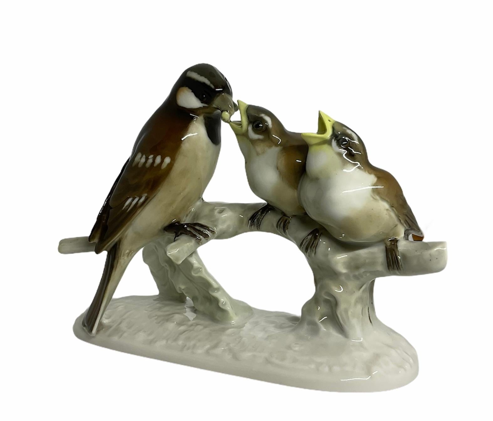 This is a Hutschenreuther porcelain group sculpture/figurine that is depicting a mother sparrow feeding one of her two baby birds. They are hand painted dark chocolate/black in the back and white in the front. The babies’ sparrows peaks are yellow.