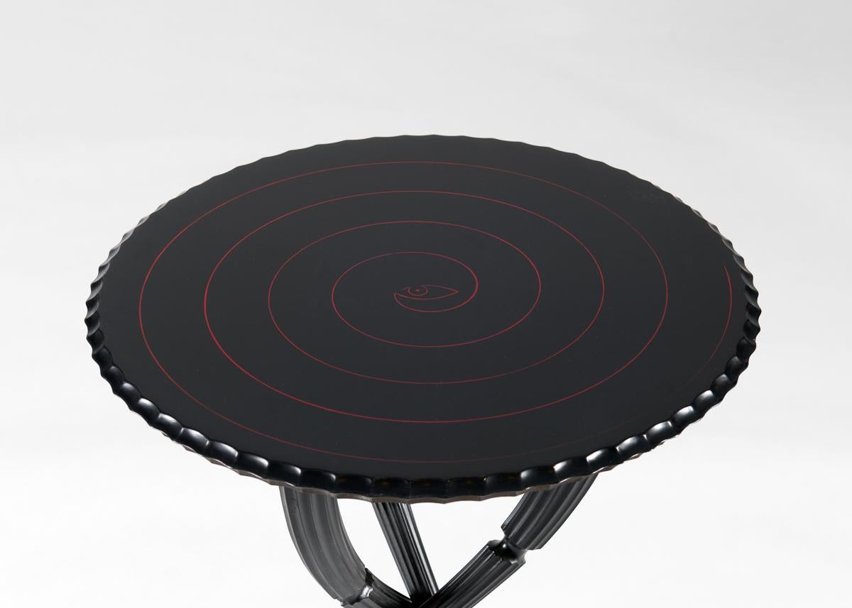 This elegant side table features a round top with a scalloped edge and rests upon three intertwining legs that taper dramatically as they descend to the floor. With it, the artist has assembled forms which do not merely intimate motion, but draw the