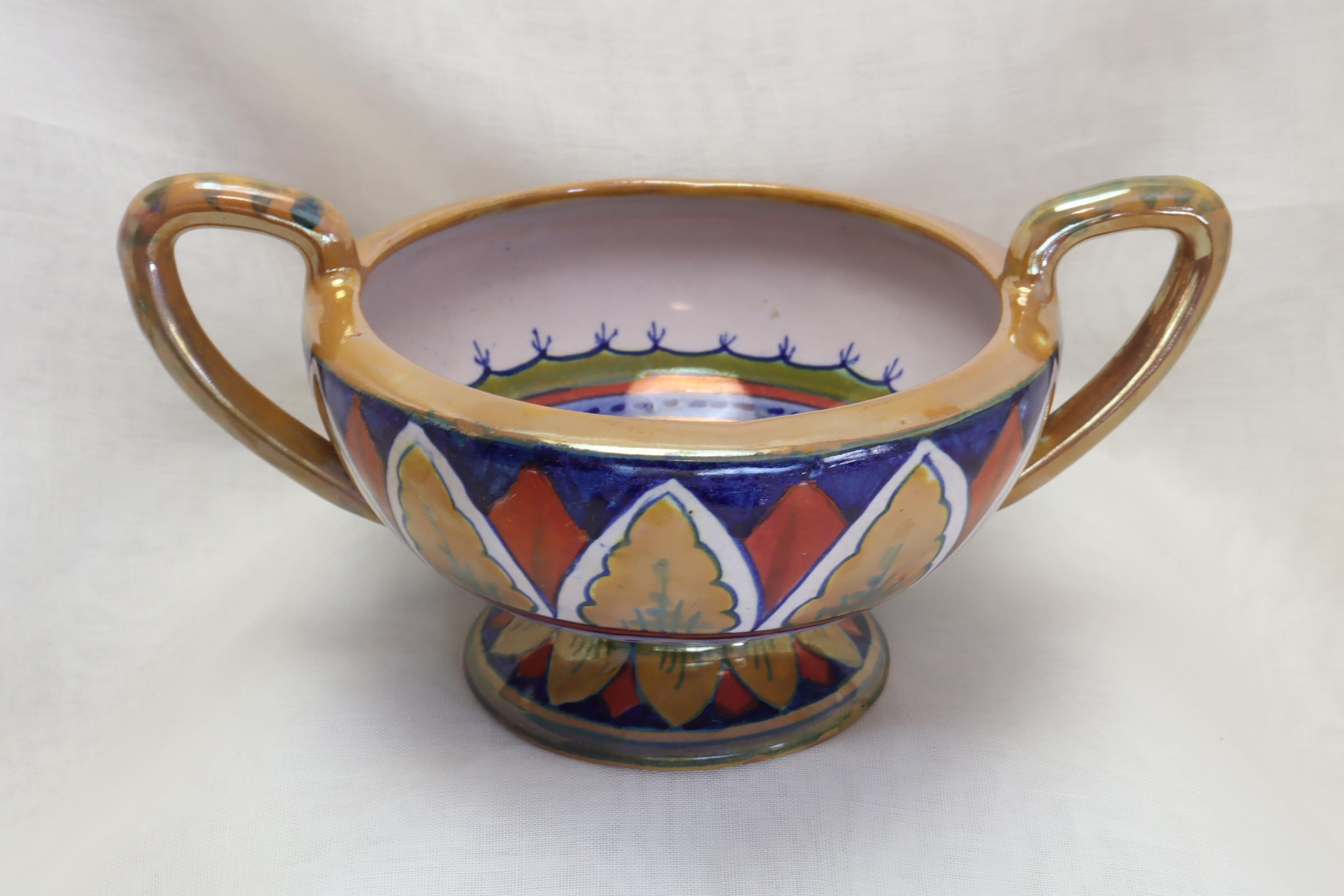 This two handled, footed bowl with a lustre glaze is by Lorenzo Rubboli of Gualdo Tadino in Umbria, Italy. The centre of the bowl is decorated with a stylised flower which is contained within several differently coloured rings. The outside features