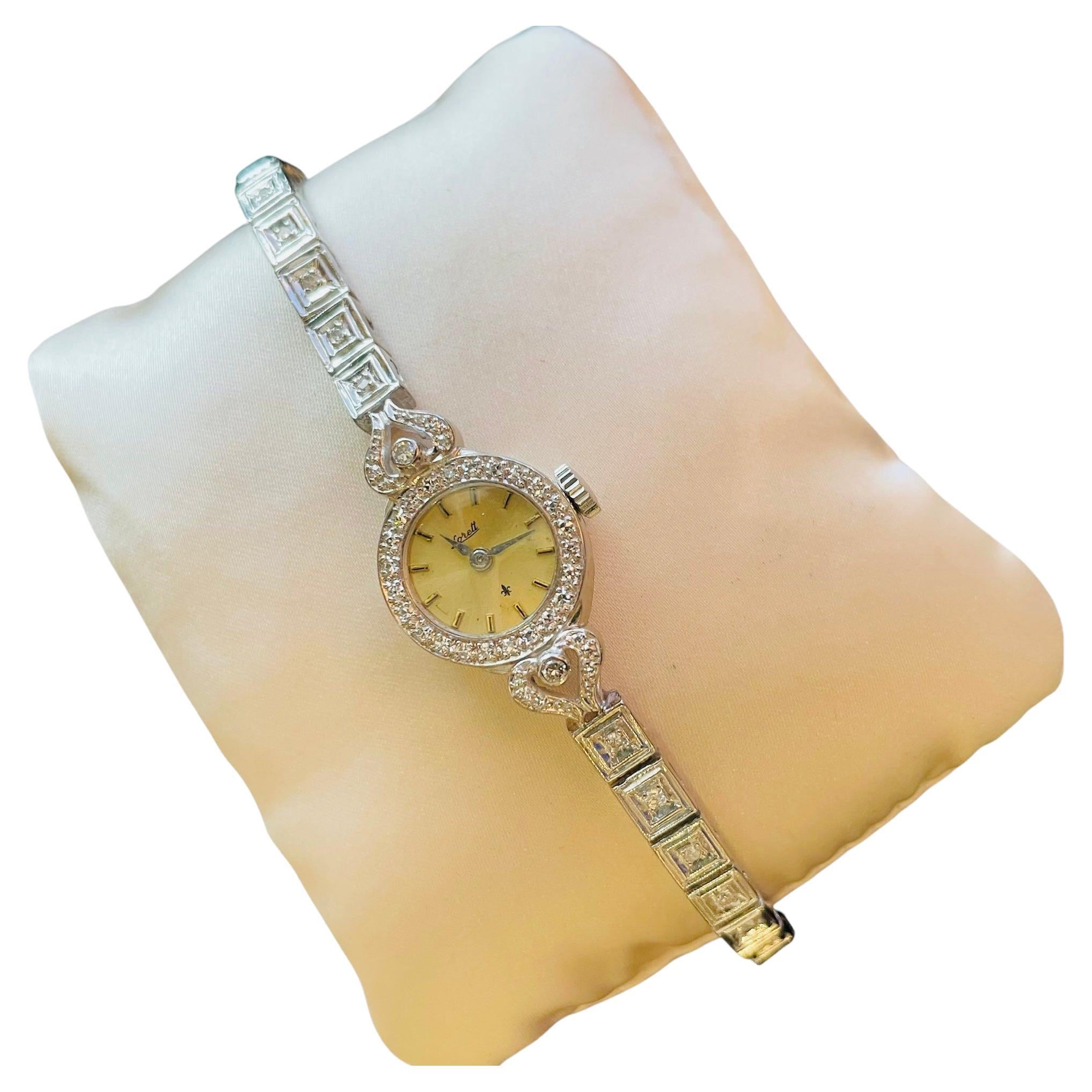 This is a Lorett Corletto Lady’s Wrist Watch. It depicts an 18K/750 white gold round case with beige color dial that has stick “numerals” and color hour/minute hands. An acrylic material glass covers the case. The case is decorated by a halo of 18k