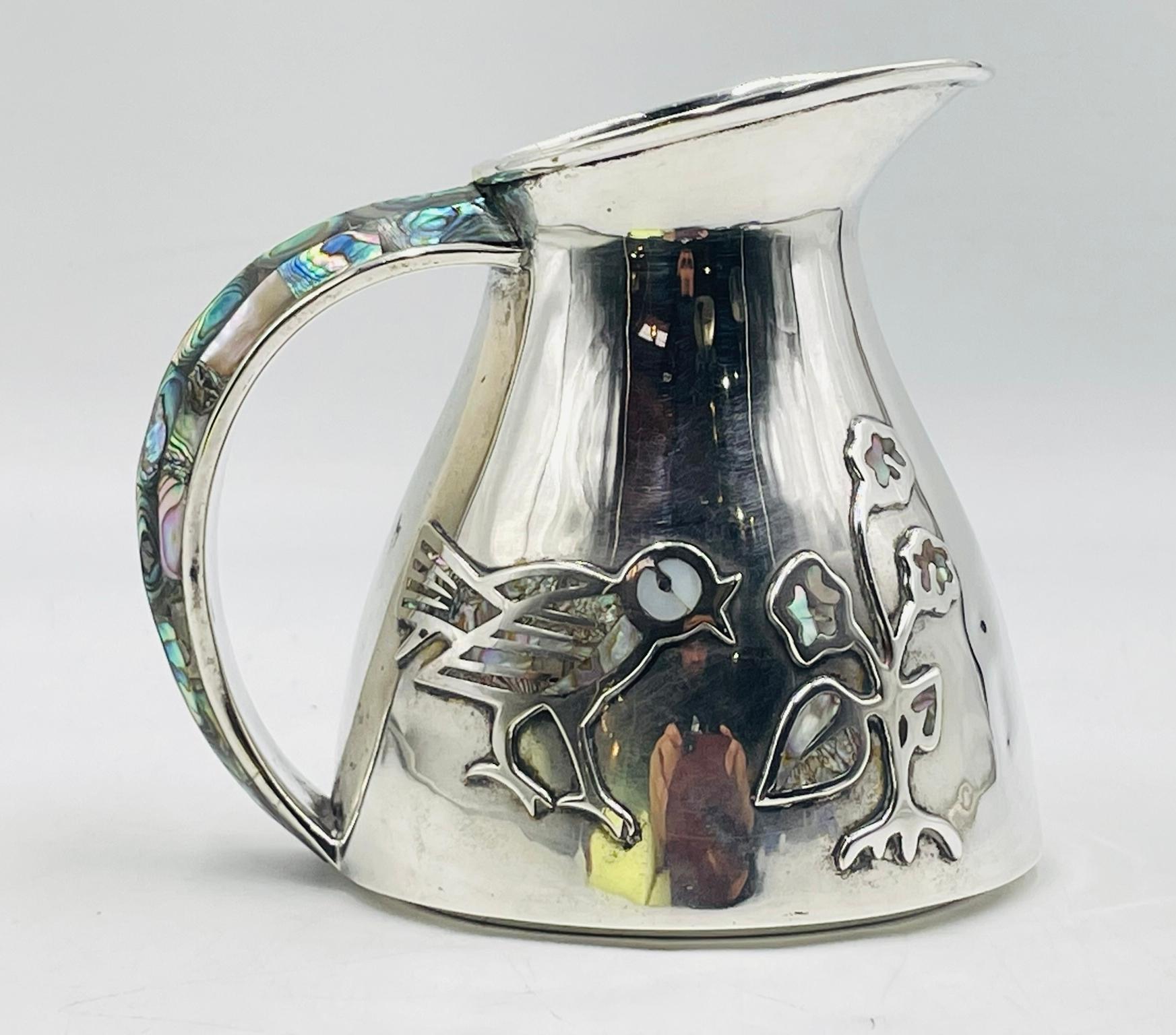 Ein wunderschön gearbeiteter, versilberter Krug von Los Castillo, einer der berühmtesten Silberschmiedefamilien Taxcos. Dieses Stück zeigt stilisierte ausgeschnittene Vogel- und Blumenmotive, die mit schimmerndem Abalone und Perlmutt eingelegt sind