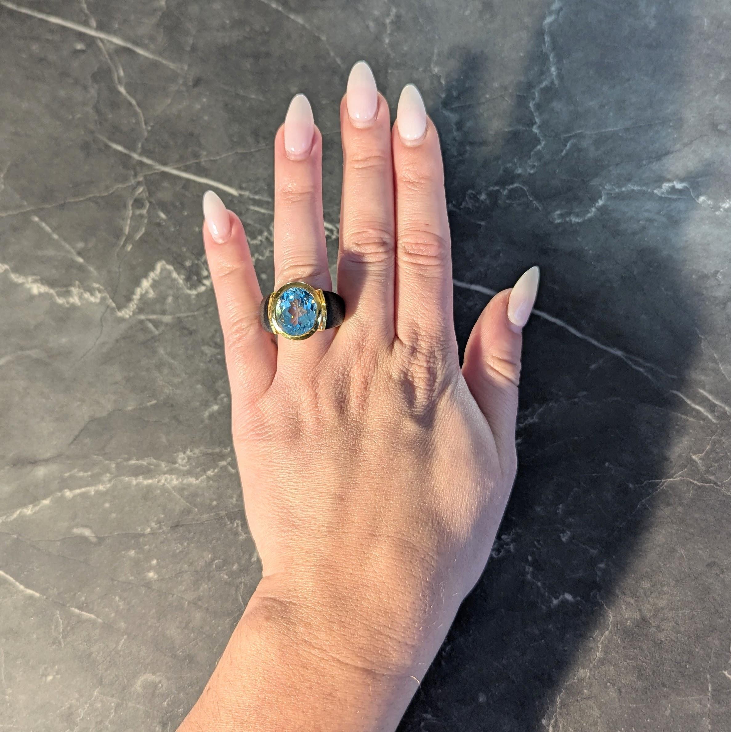 Centering an oval cut topaz weighing approximately 11.95 carats
Transparent vivid blue color and set in a high polish gold bezel
With an open floating illusion profile flanked wood shoulders
Carved dark brown in color with visible grain