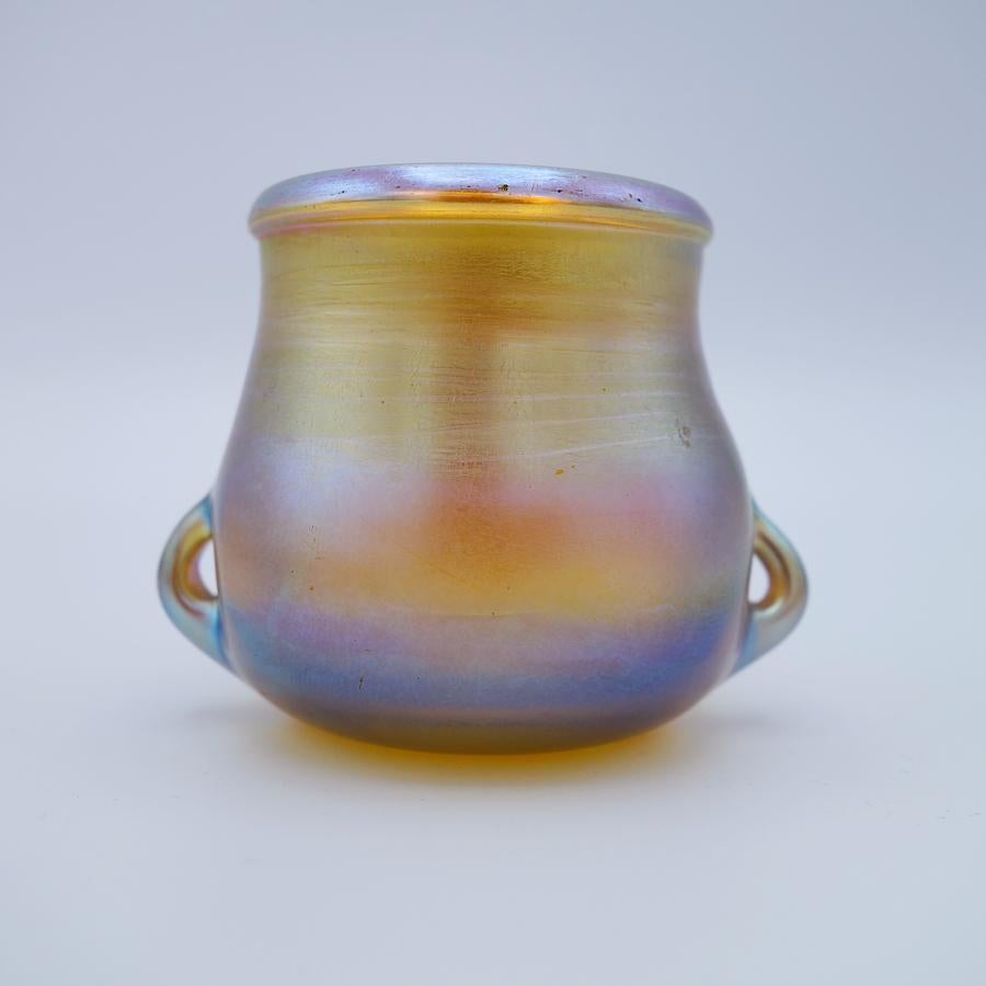 Offering this diminutive Louis Comfort Tiffany gold Favrile iridescent art glass cabinet vase. This vase features a tapered and slightly cinched cylindrical body with rounded bottom flanked by two small pulled handle designs. Signed on the