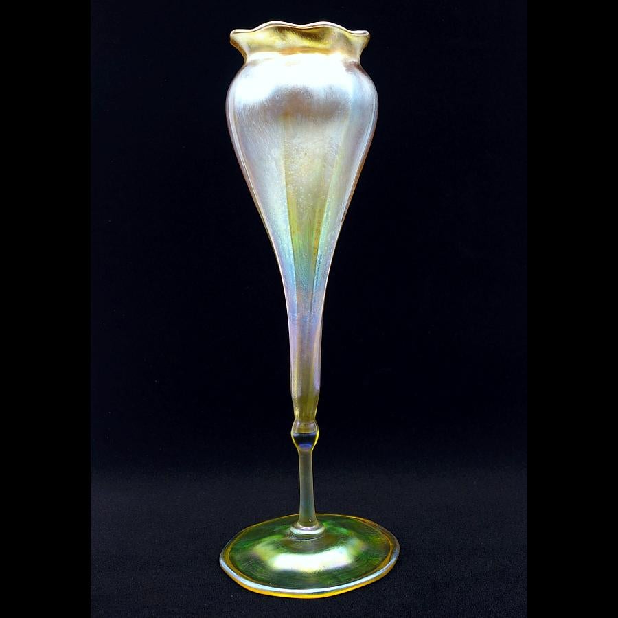 Offering this delightful Louis Comfort Tiffany gold Favrile iridescent art glass footed floriform vase. This vase features a tapered conical body with a thin stem on a round foot base and features a scalloped lip. Signed on the underneath 