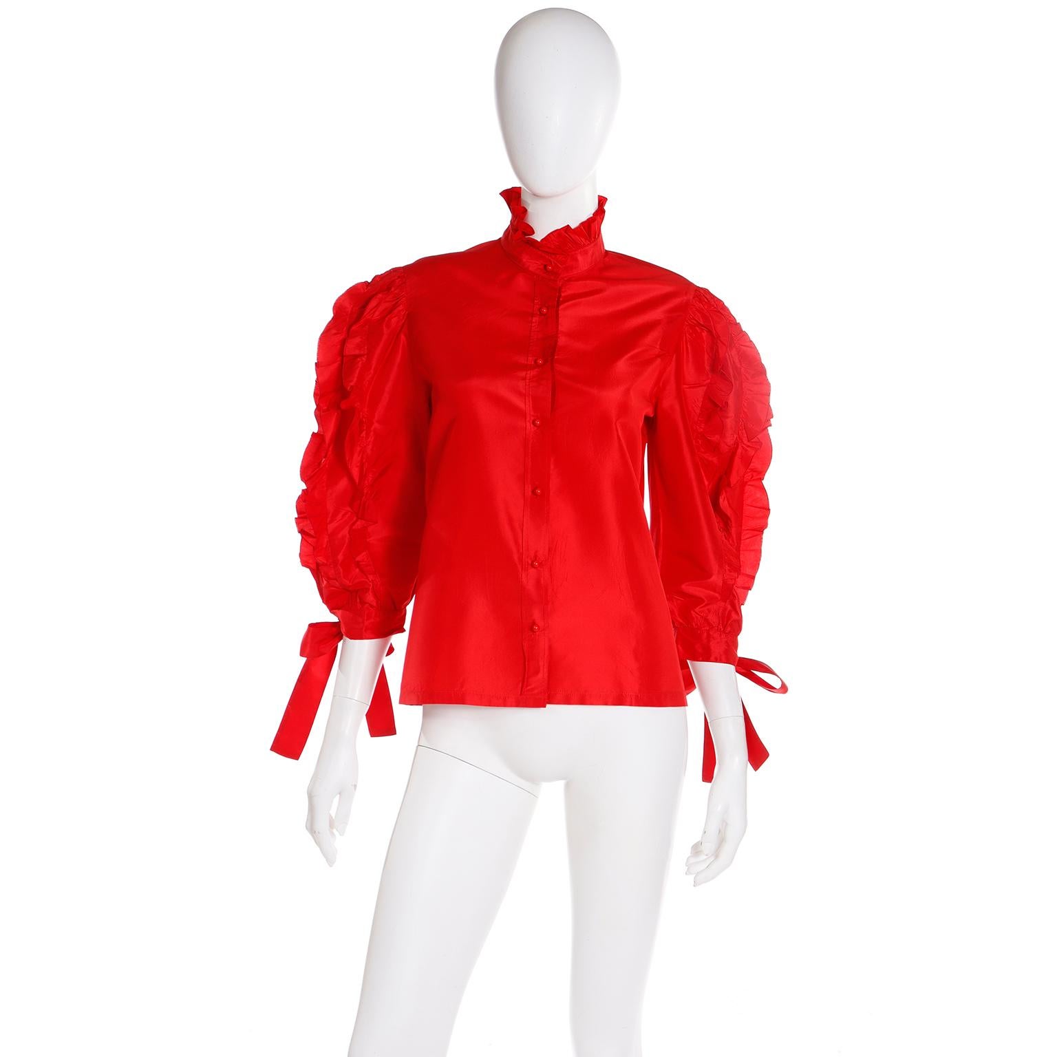 Ce chemisier Louis Feraud vintage des années 1980 est en satin de soie rouge et présente de jolis volants et liens. Ce haut a juste ce qu'il faut de dramatique avec un col à volants et des boutons boules rouges au centre du devant. Chaque manche a