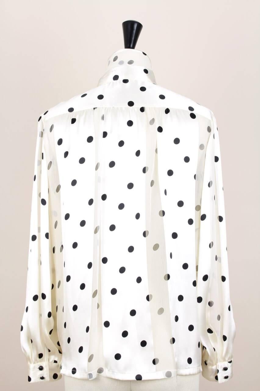 This is a wonderful semi-transparent polka dot silk blouse by Louis Féraud from the 1980s. It is made from alternating high quality silk satin and silk chiffon in a cream hue with black polka dots. The blouse features a high neck with a tie that is