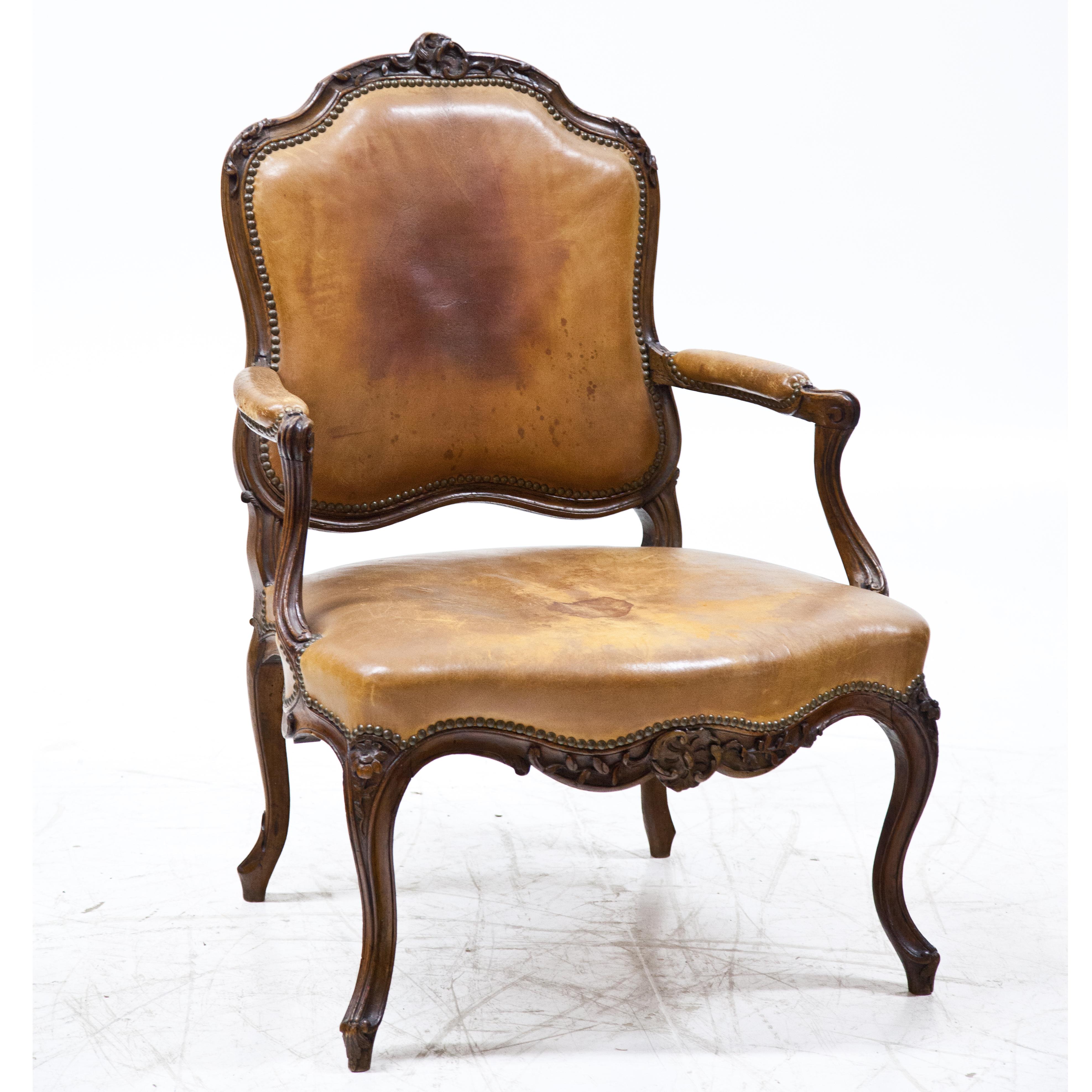 Armchair standing on S-legs with curved frame and carved floral decorations. The trapezoidal seat as well as the back and armrests are covered with riveted leather. The slightly set back armrests rest on curved, rocaille-shaped supports. The curved