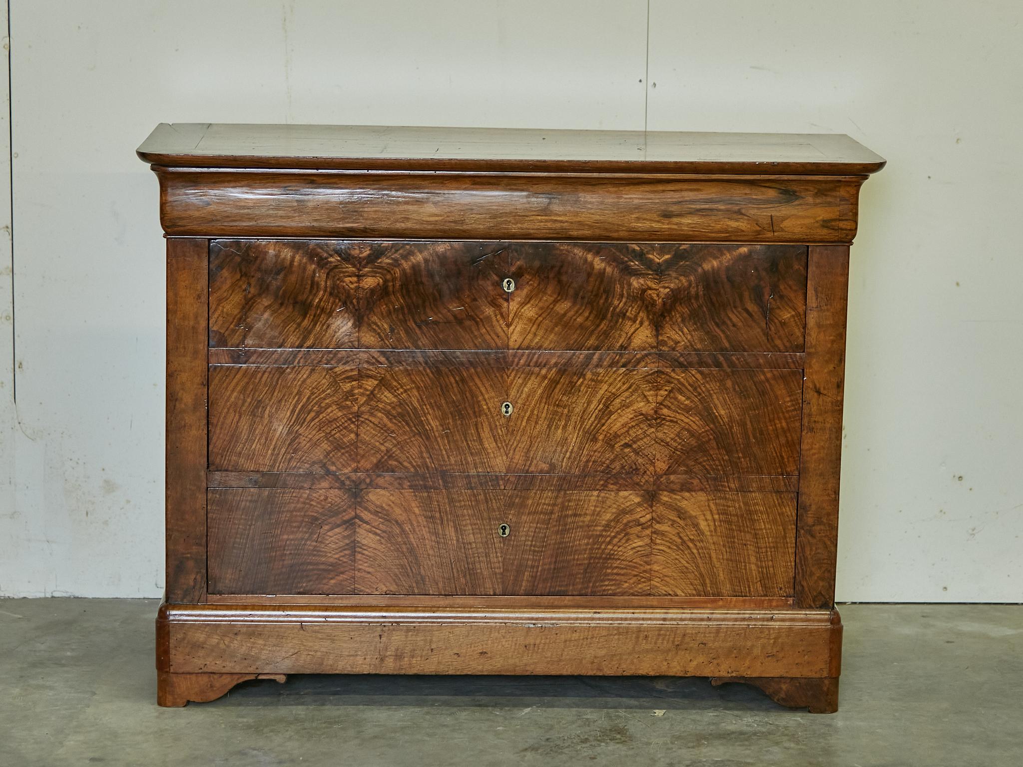 A French Louis-Philippe walnut four-drawer commode from the 19th century with bookmatched façade and bracket feet. Step into the realm of classic French elegance with this French Louis-Philippe walnut four-drawer commode from the 19th century, a