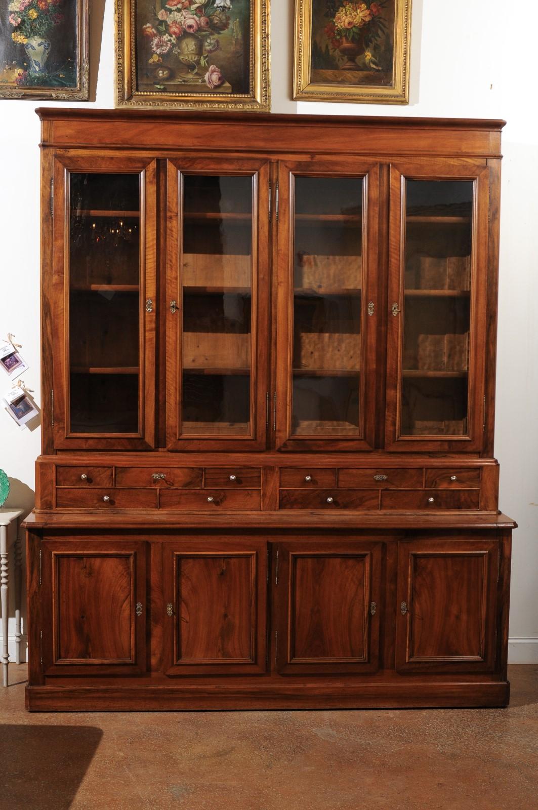 A French Louis-Philippe style walnut bookcase from the late 19th century with glass doors, drawers and wooden doors. This French two-part walnut bibliothèque features a molded cornice sitting above four glass doors opening to reveal adjustable