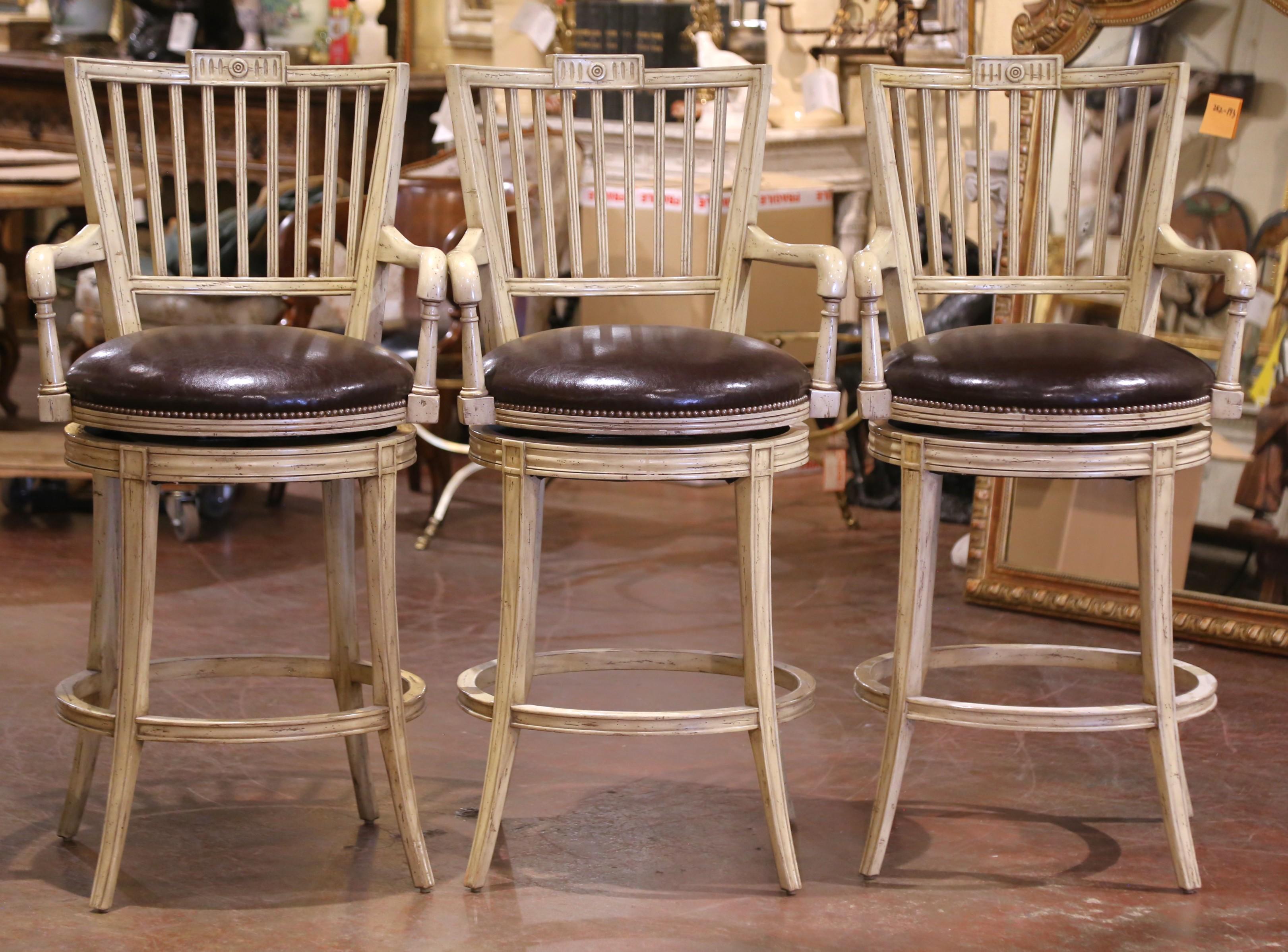 Offered as a set of three, these comfortable and stylish French barstools were crafted circa 1990 and feature a smooth 360° swivel mechanism, blending practical function with timeless design. The cream-painted wood frames have a gently distressed