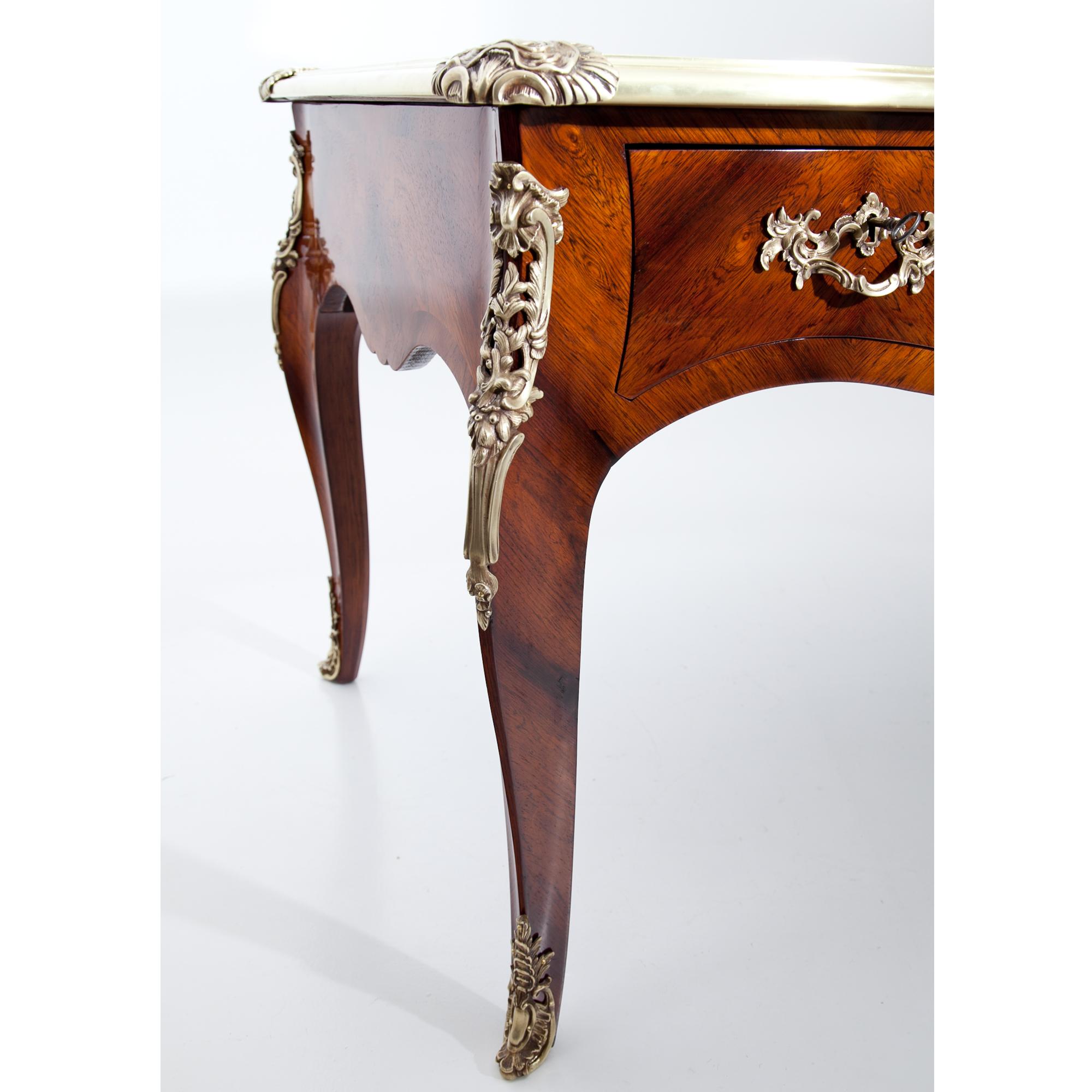 Large Louis-Quinze-style desk in mahogany veneered. The writing surface is covered in brown leather, the edges are decorated with brass profiles. The desk has three drawers, which are imitated on the back. The curved legs show decorative fittings in