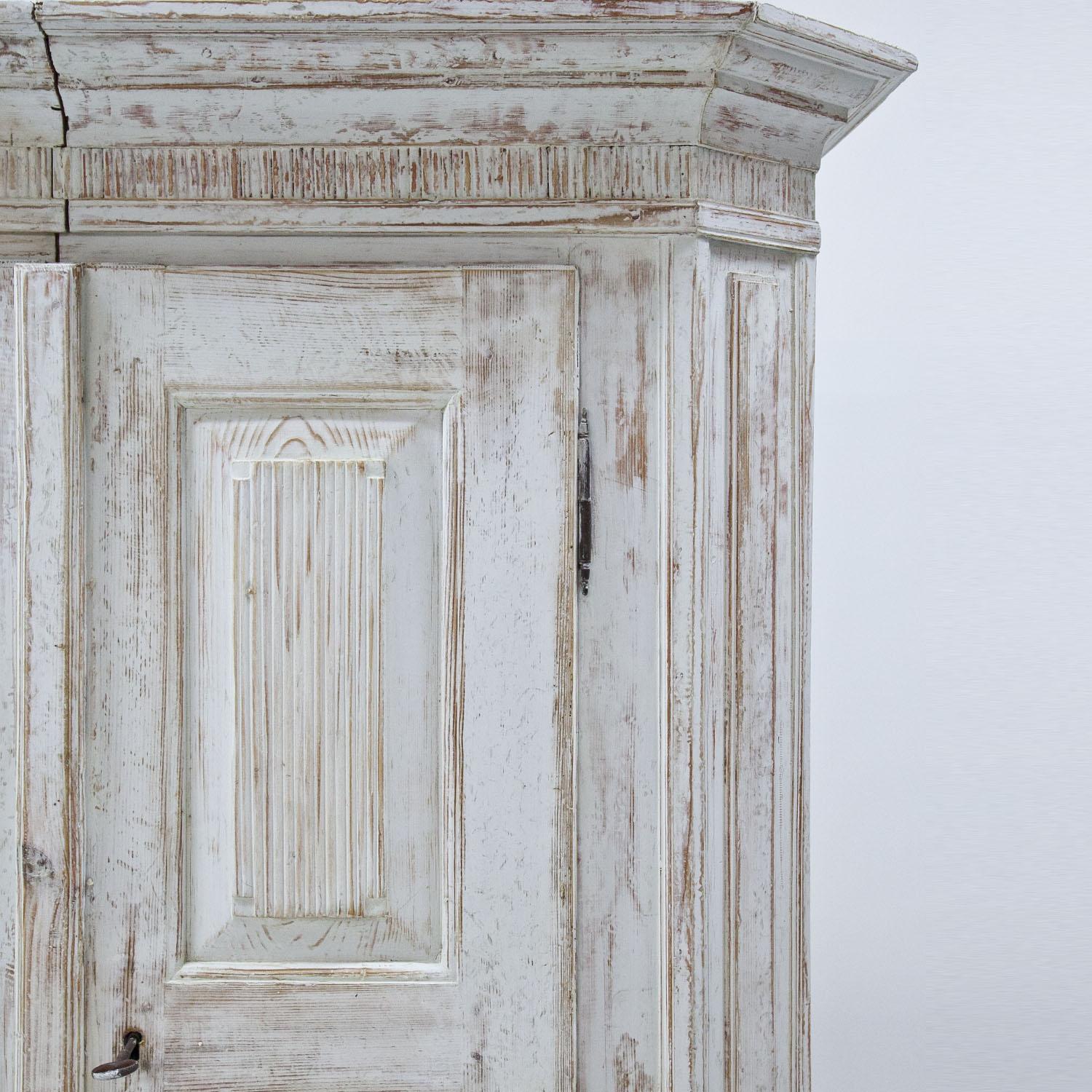 Two-doored armoire on short feet. The body with slanted edges, a protruding cornice and fillings on the doors, the base and the sides. Decorative vertical grooves on the doors and below the cornice. The white paint is new and has a worn-looking