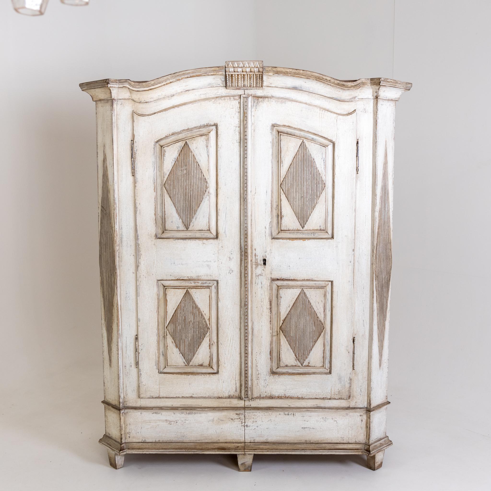 Two-door cabinet with a wide plinth zone and curved cornice. The corpus is decorated with fluted diamond patterns and filling fields on the doors. The creamy white frame is new and has been decoratively patinated.