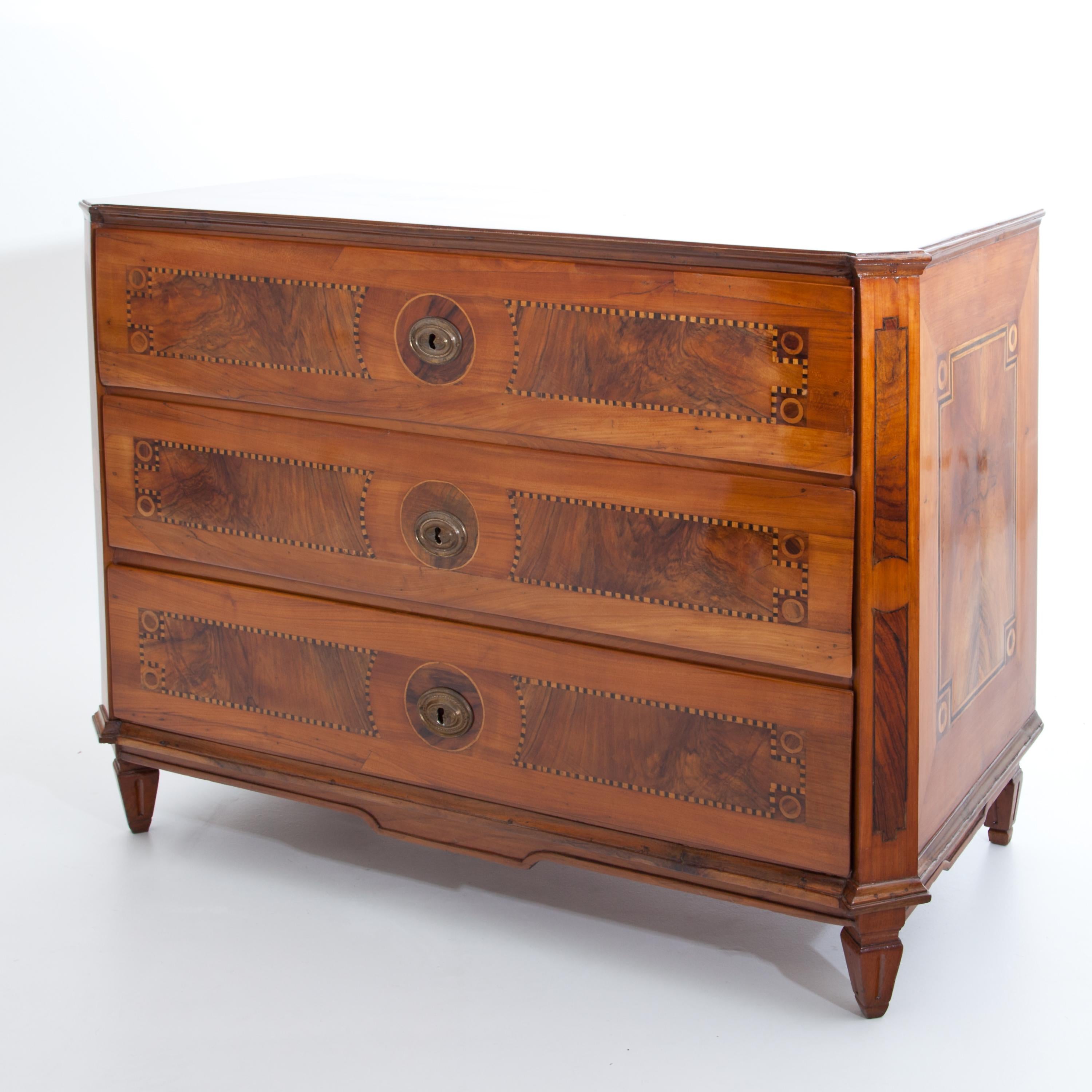 A three-drawered chest of drawers made of cherry on low fluted square pointed feet, with bevelled, slightly cranked corners. The body is inlaid with burl wood in the form of fillings and framed by fillet bands, the top is additionally decorated with