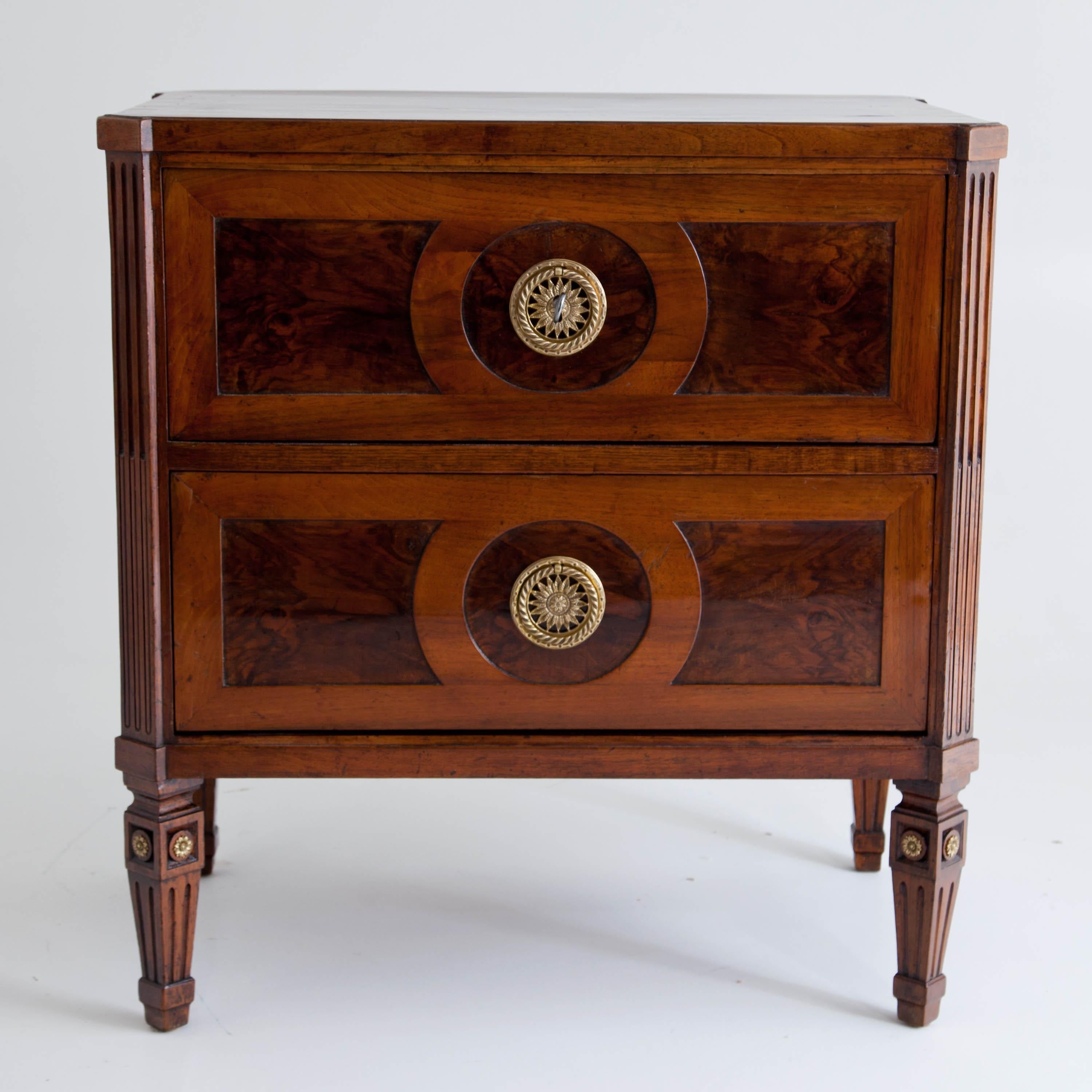 Small two-drawered chest of drawers on fluted square pointed feet with fire-gilt rosette decoration. The bevelled pilaster-shaped corners show fluting and pipes. Filling panels on the front and sides in walnut root wood. The fronts of the drawers