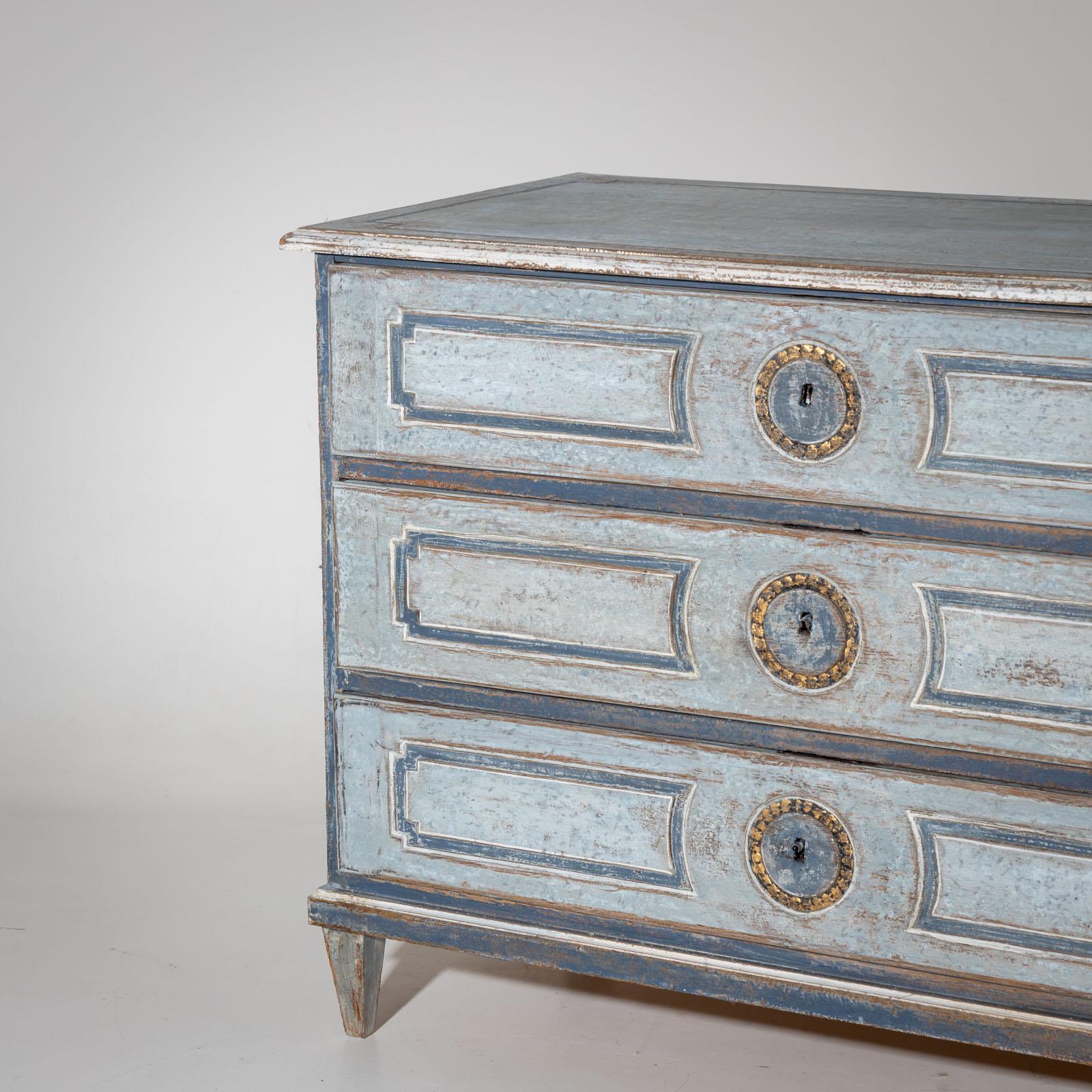 Hand-painted, blue chest of drawers with three drawers. Panels adorn the front and sides. The keyholes are set off with a gold-patinated ring. The chest of drawers has been repainted and given an antique patina.