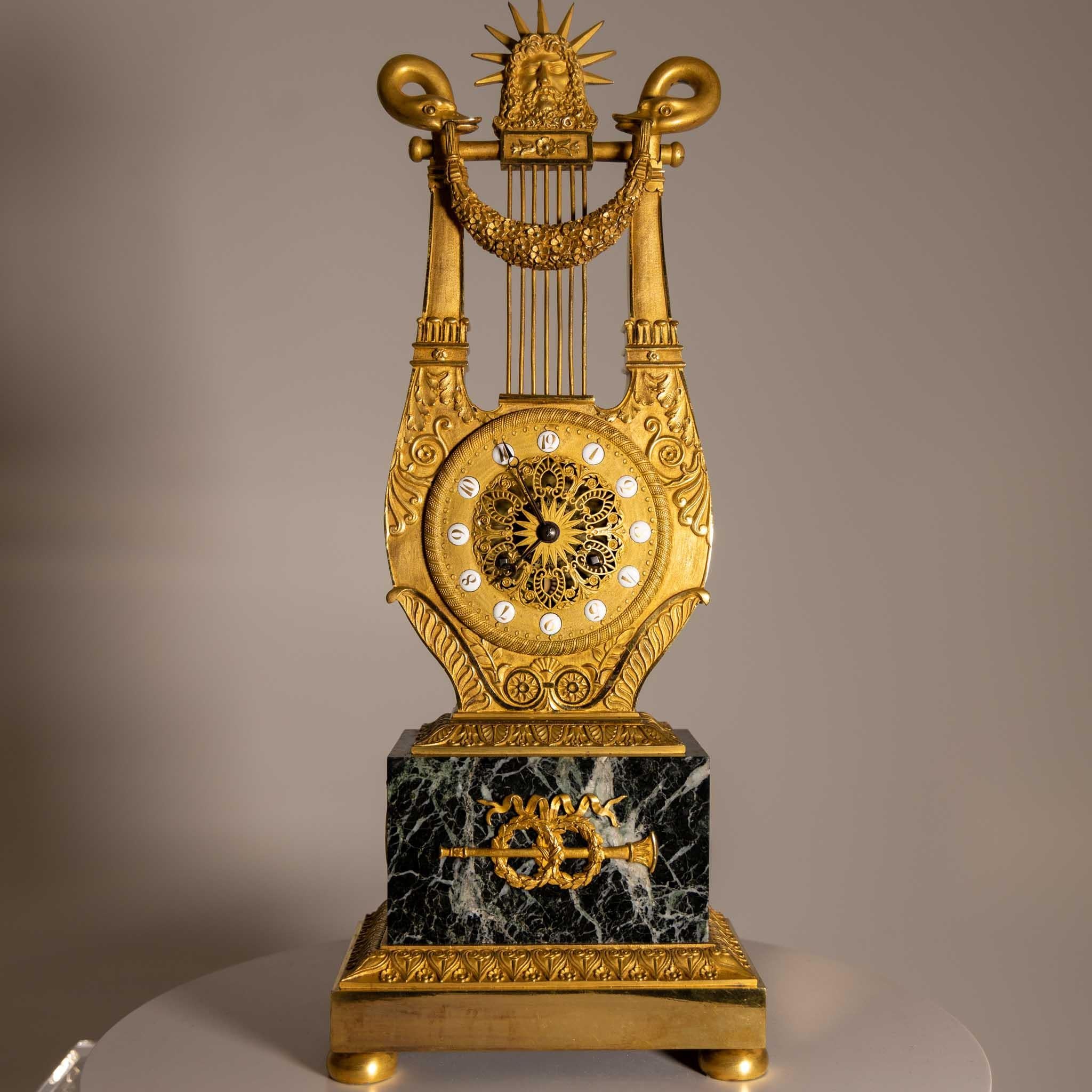 Lyre-shaped mantel clock on marble base with crowning head of sun god Apollo and swan heads holding a flower festoon in their beaks. The dial is decorated with round enamel numerals. Fire-gilded bronze. Type illustrated in: Elke Niehüser: French