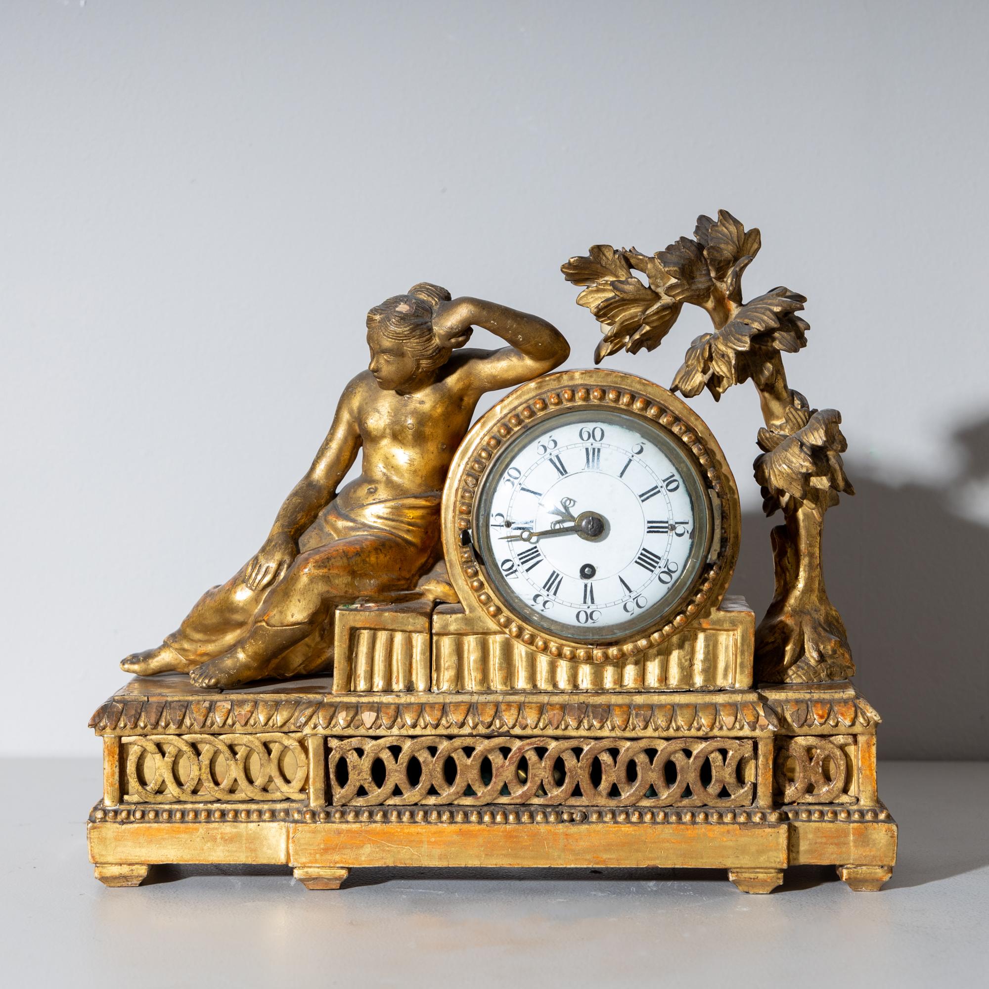Louis Seize mantel clock or pendule, housed in a gold-patinated wooden case featuring intricate openwork rings and a round clock case. The design depicts a young undressed woman leaning against it, with a tree on the right side providing shade. The