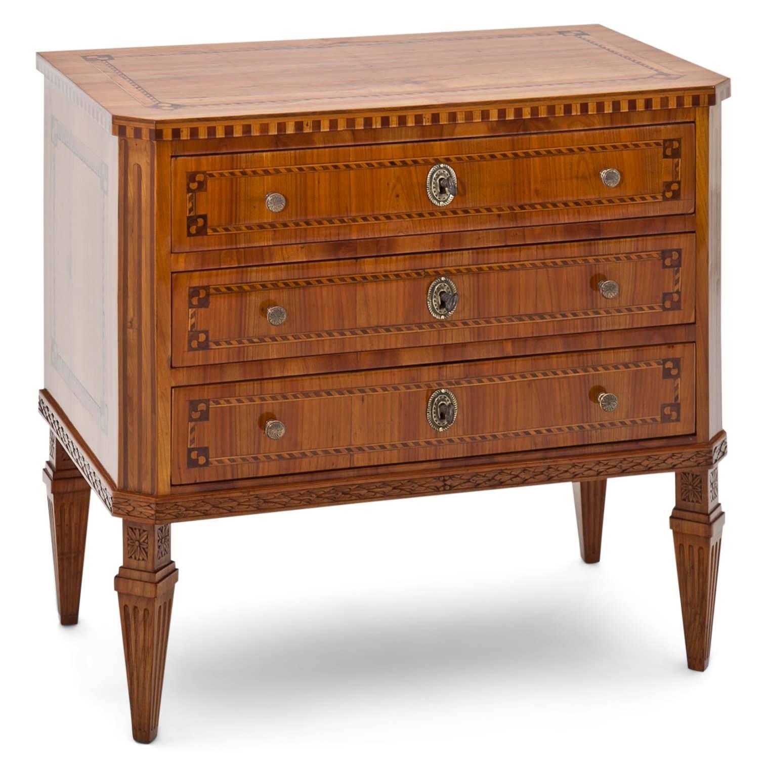Three-drawered Louis Seize-style chest of drawers on fluted tapered feet with carved flower ornaments. The body with slanted edges and inlays on front, sides and on top. The fittings are new. This chest is a Louis Seize-style chest, designed under