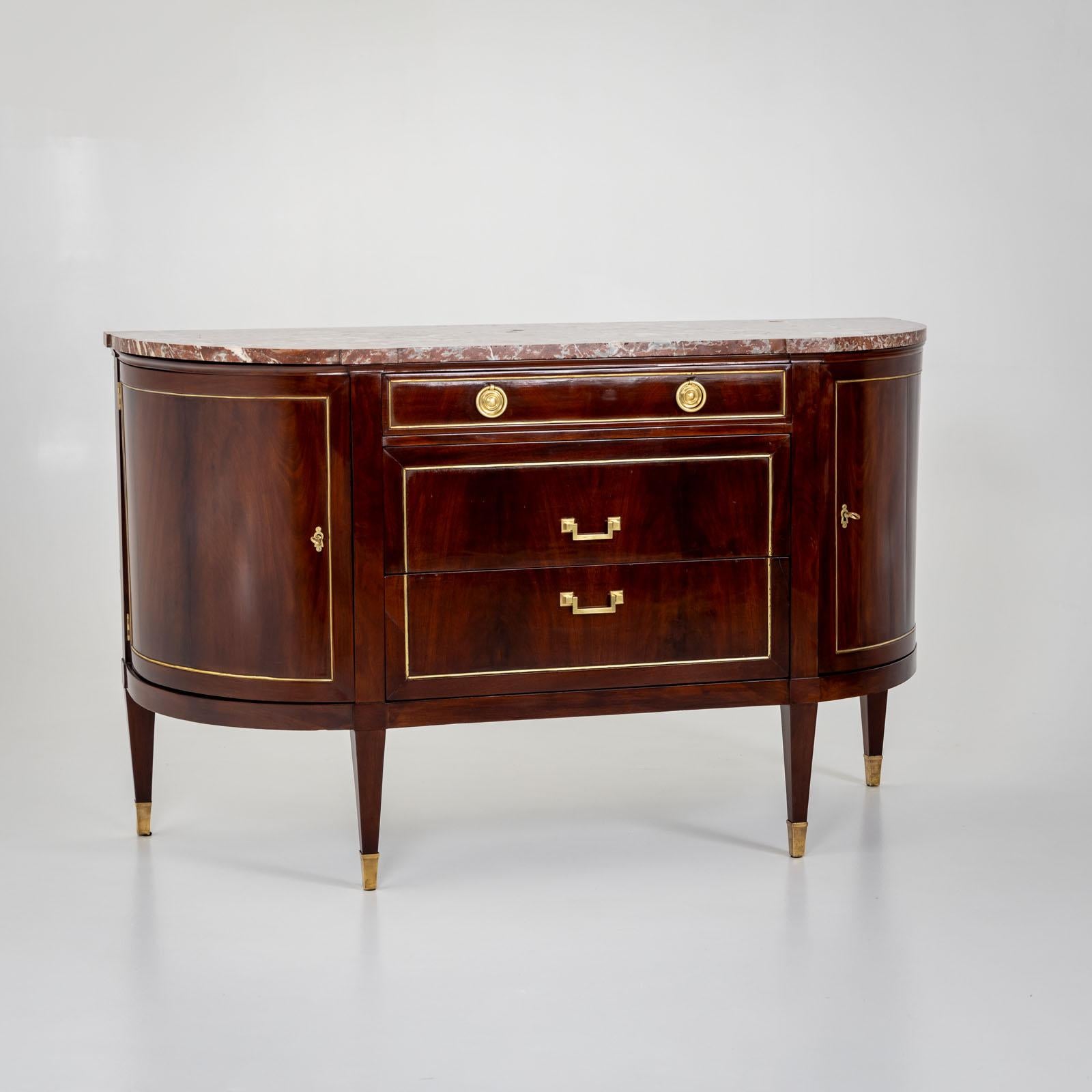 Demi-lune sideboard in Louis Seize style with red and white marble top and two doors and three drawers. The doors are framed with brass profiles. The sideboard is veneered in mahogany and stands on square tapered feet with brass sabots. 