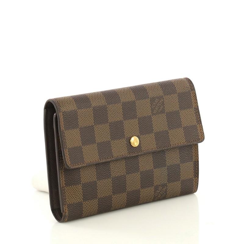This Louis Vuitton Alexandra Wallet Damier, crafted from damier ebene coated canvas, features leather trim and gold-tone hardware. Its snap button closure opens to a brown leather interior with multiple card slots, snap pocket, and slip pockets.