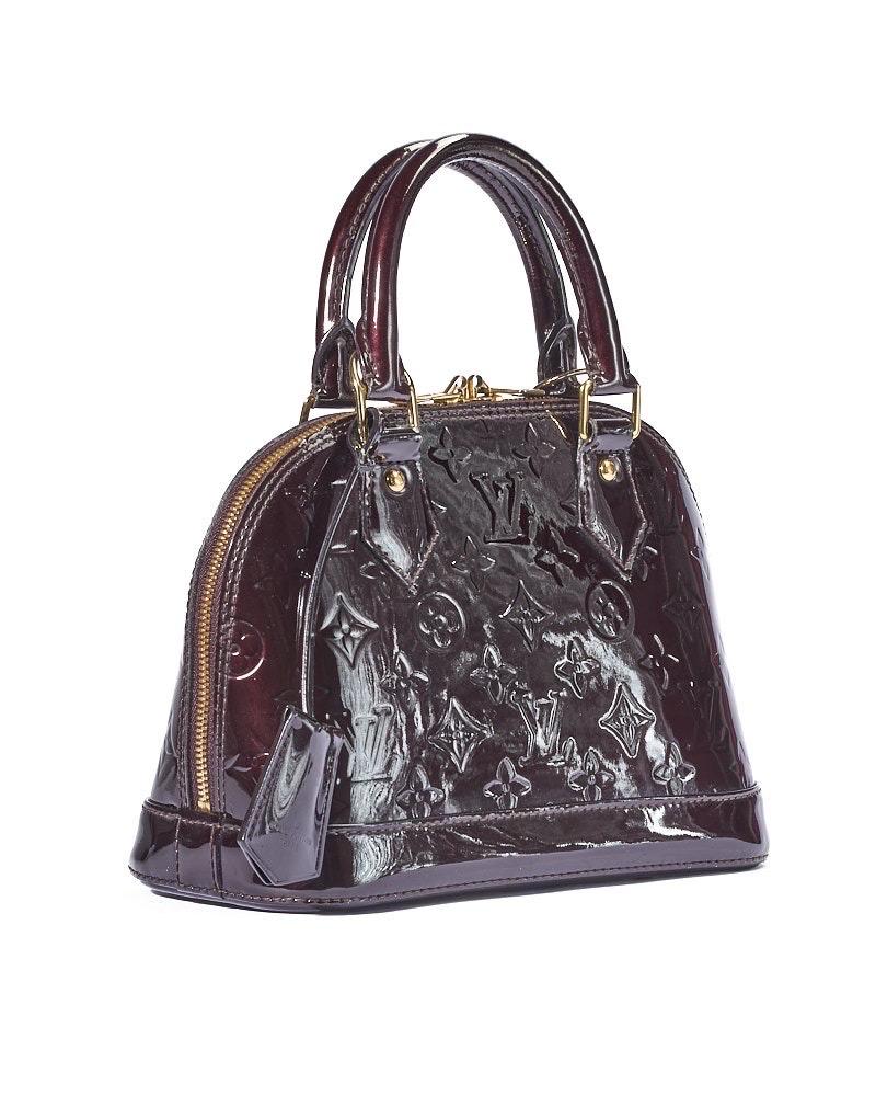 Borsa firmata Louis Vuitton, modello Alma nella misura BB, realizzata in pelle verniciata nella colorazione amaranto con motivo monogram e hardware dorato. Dotata di una chiusura semicircolare a zip con doppio cursore, internamente rivestita in tela