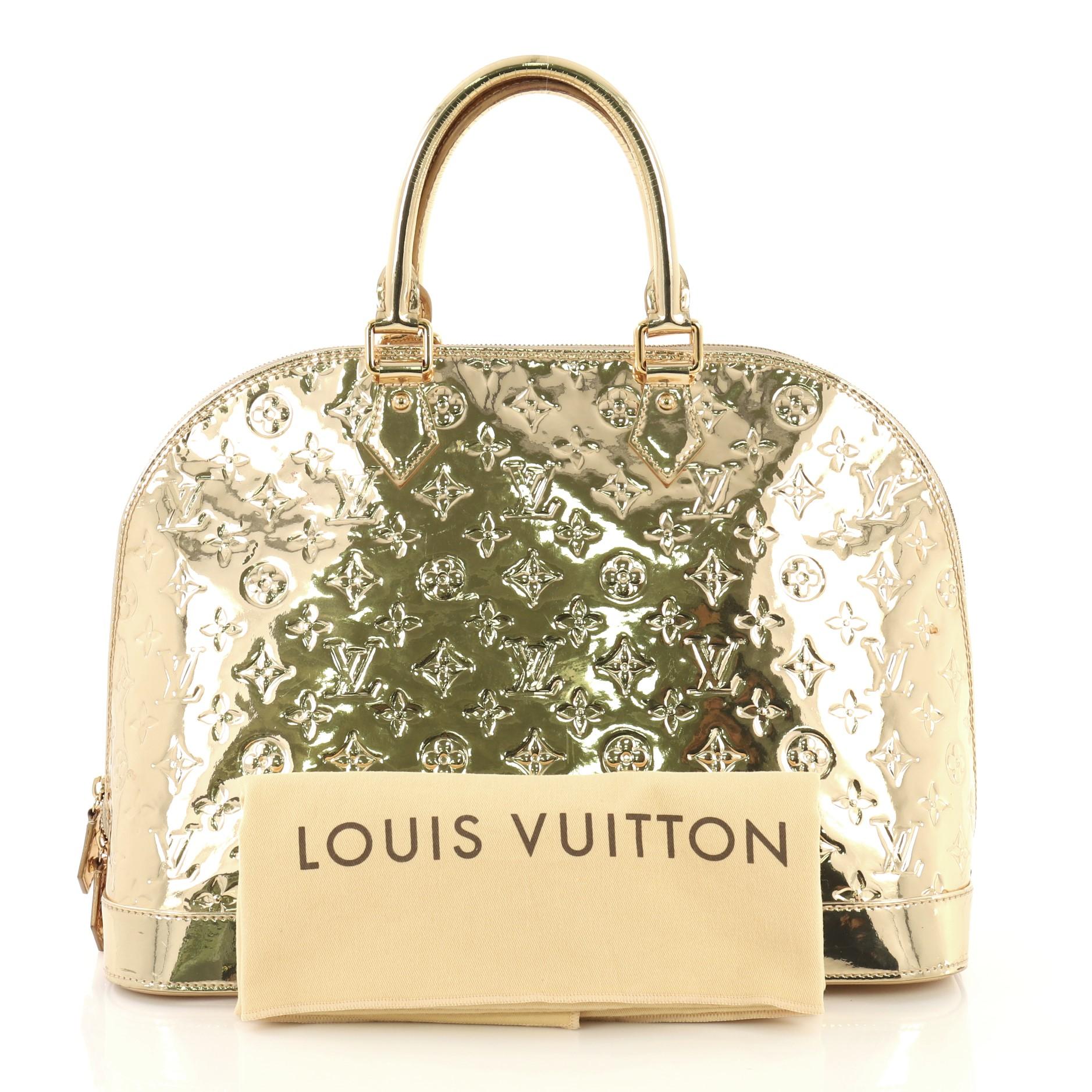 This Louis Vuitton Alma Handbag Miroir PVC GM, crafted from gold miroir PVC, features dual rolled handles, protective base studs, and gold-tone hardware. Its zip closure opens to a neutral fabric interior with slip pockets. Authenticity code reads:
