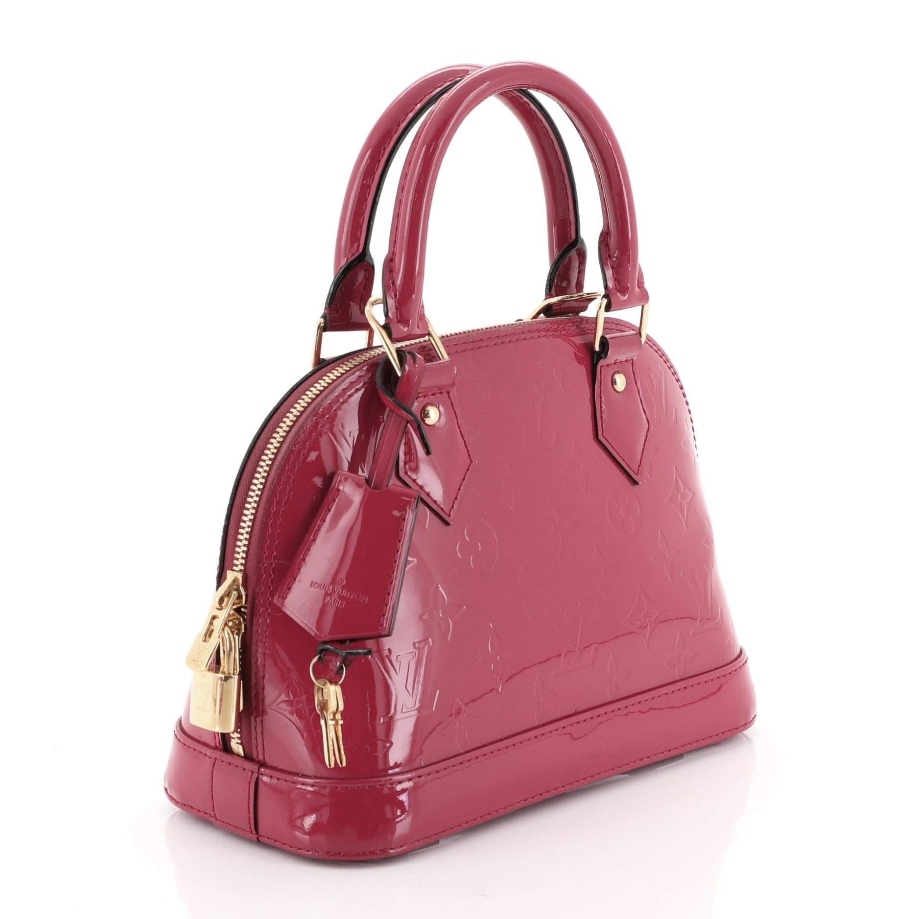 This Louis Vuitton Alma Handbag Monogram Vernis BB, crafted from pink monogram vernis leather, features dual rolled handles, protective base studs, and gold-tone hardware. Its two-way zip closure opens to a pink fabric interior with slip pocket.