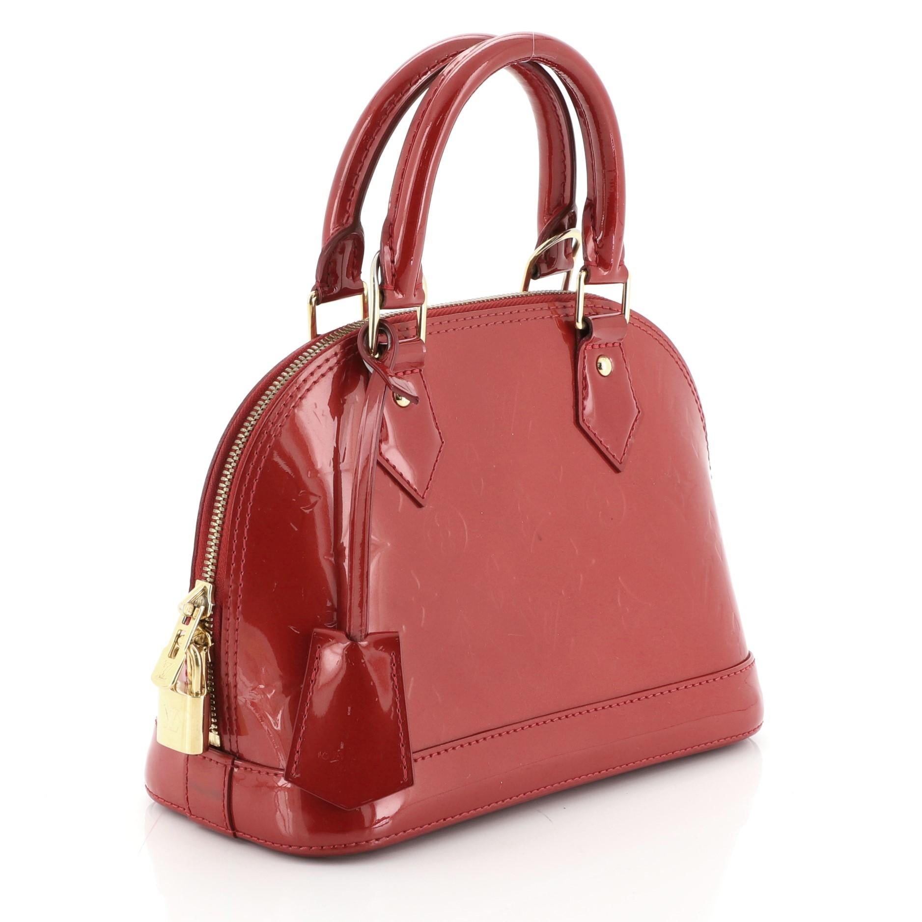 This Louis Vuitton Alma Handbag Monogram Vernis BB, crafted from red monogram vernis leather, features dual rolled handles, protective base studs, and gold-tone hardware. Its two-way zip closure opens to a red fabric interior with slip pocket.