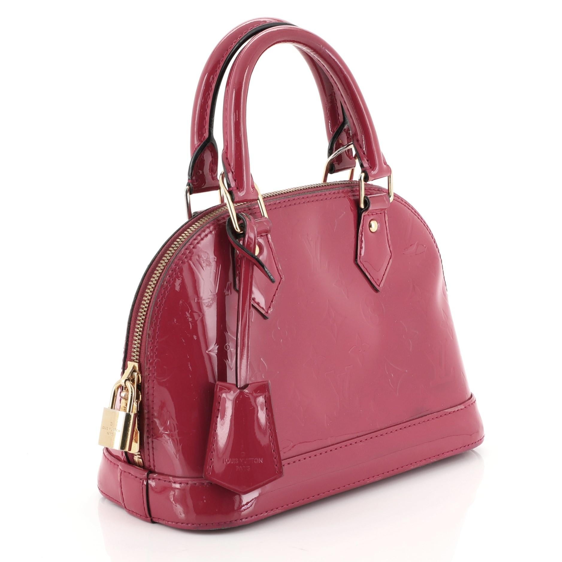 This Louis Vuitton Alma Handbag Monogram Vernis BB, crafted from pink monogram vernis leather, features dual rolled handles, protective base studs, and gold-tone hardware. Its two-way zip closure opens to a pink fabric interior with slip pocket.