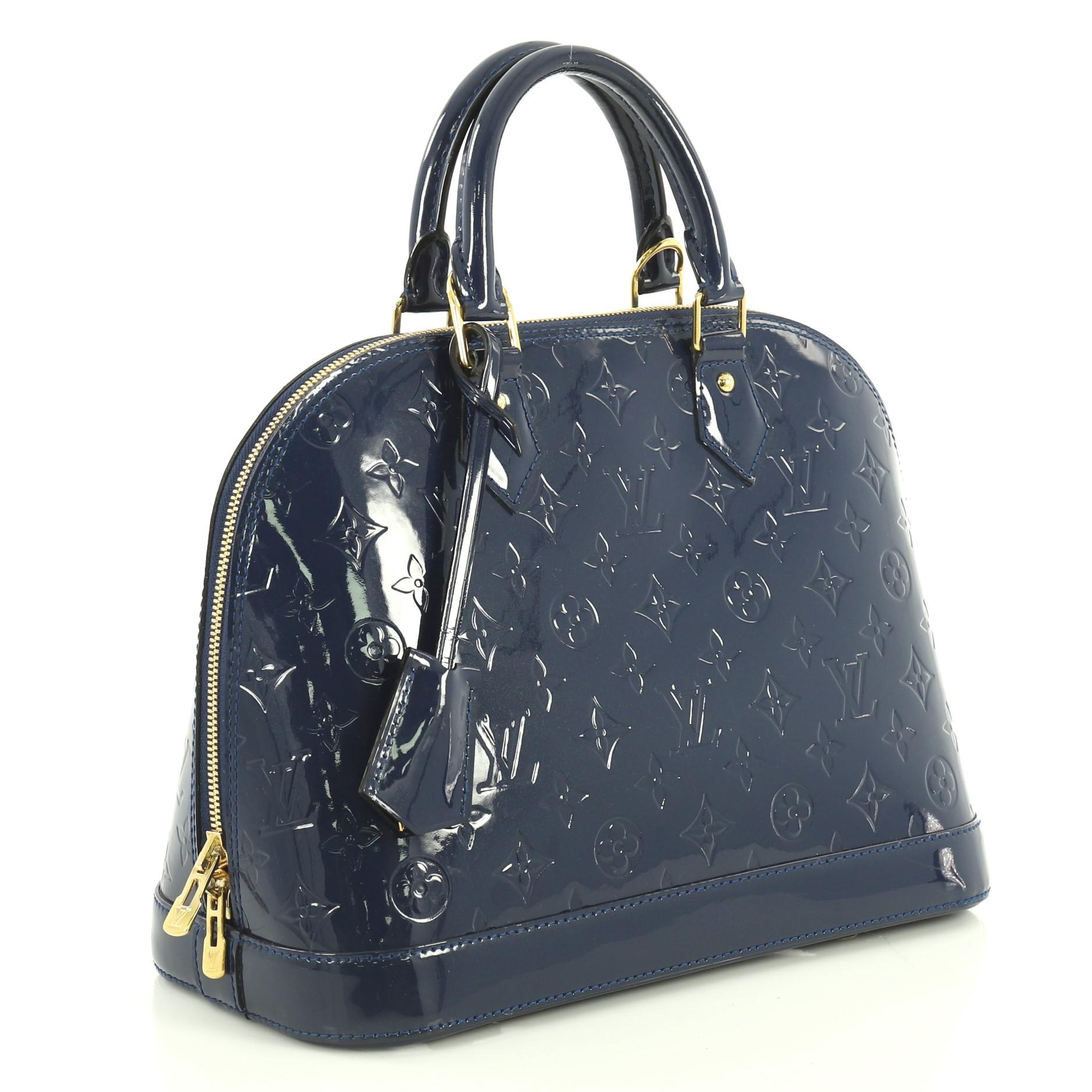 This Louis Vuitton Alma Handbag Monogram Vernis PM, crafted from blue monogram vernis leather, features dual rolled handles, protective base studs, and gold-tone hardware. Its zip closure opens to a blue fabric interior with slip pockets.