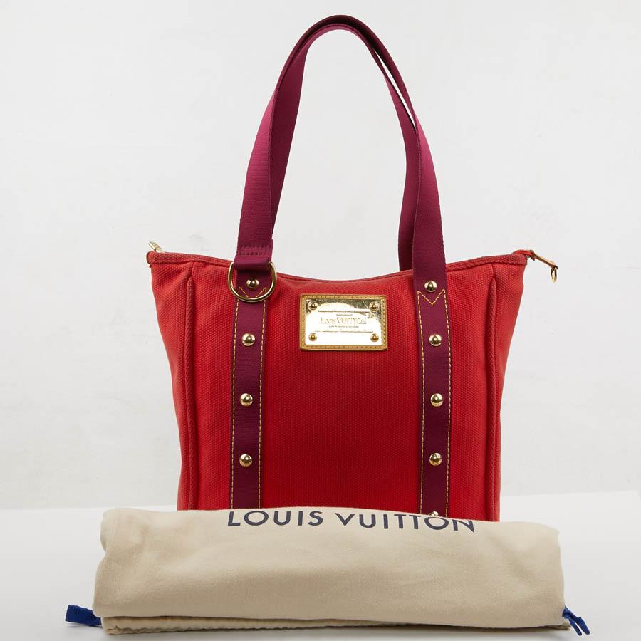 This Antigua bag from Maison Louis Vuitton is made of red cotton canvas. The two bands that serve as handles are fuchsia. It has a zipper. The metalwork is golden. The lining is in fuchsia and beige striped canvas with a slit pocket. The small